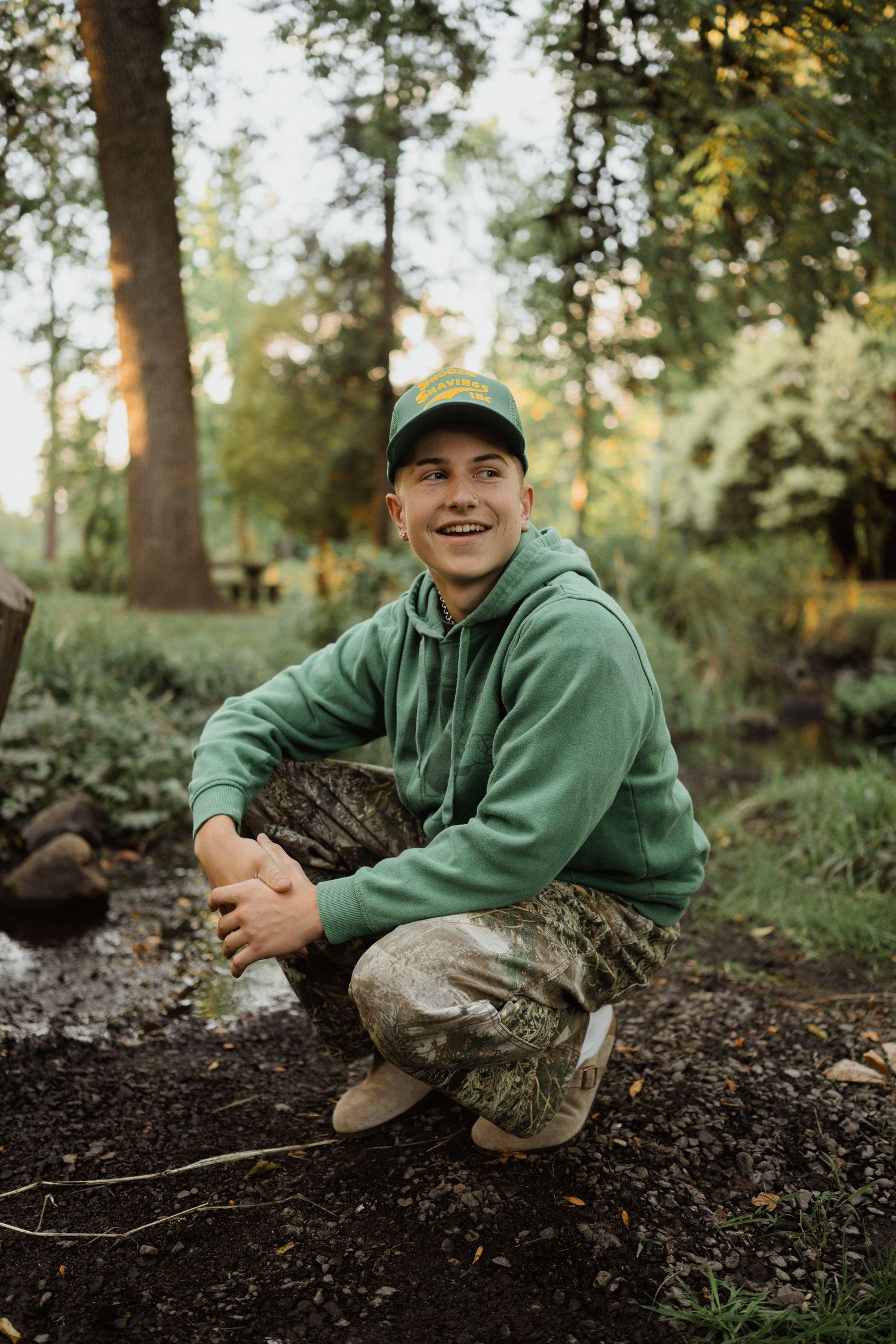 Someone wearing camo pants and a green hoodie crouches outdoors in a forest setting.