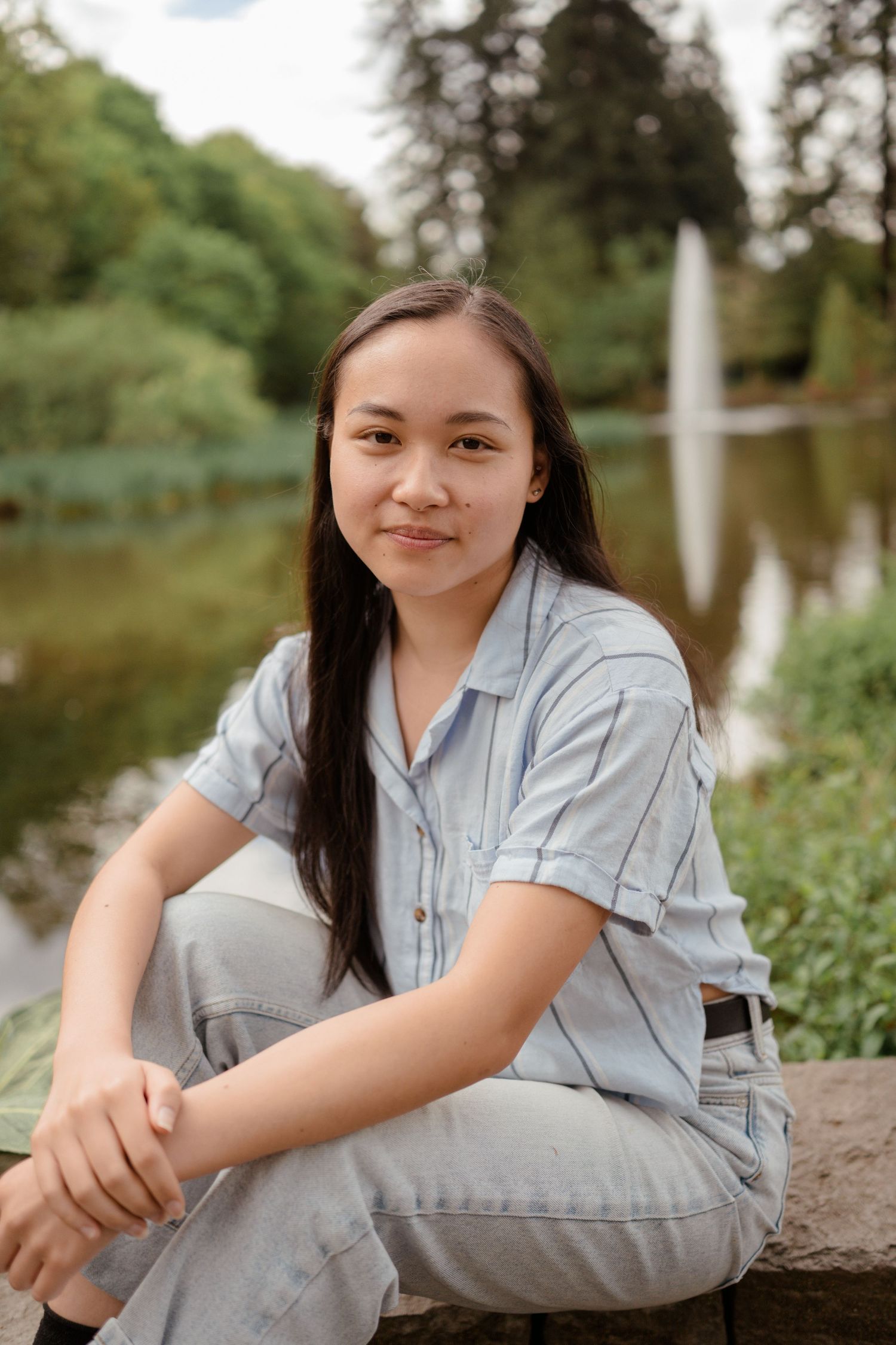 Portrait by a pond wearing a light blue shirt and casual attire while sitting on rocks.