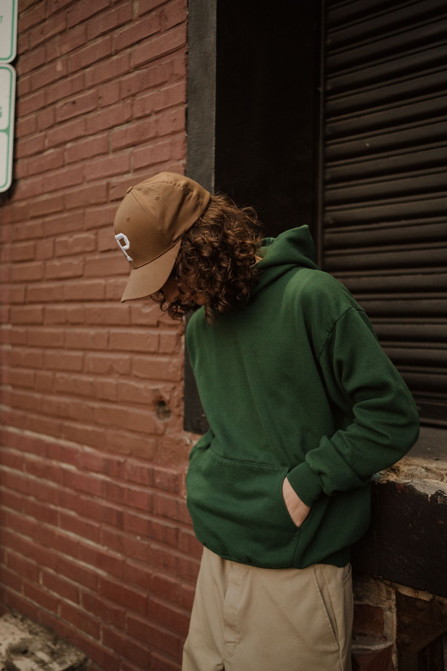 Person in green hoodie and tan cap poses against brick wall in casual urban setting.