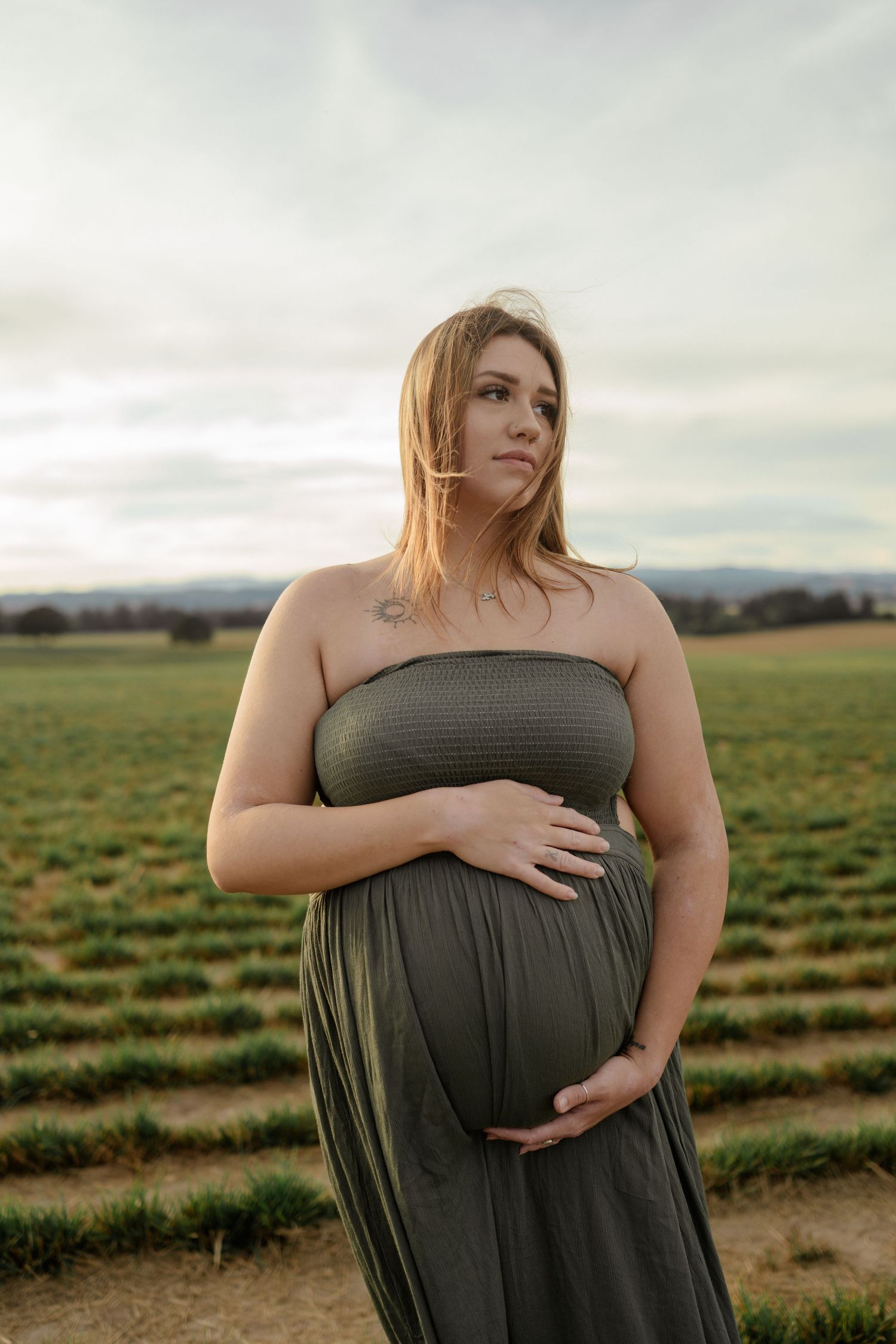 Person in gray strapless dress stands in open field during sunset placing hands on pregnant belly.