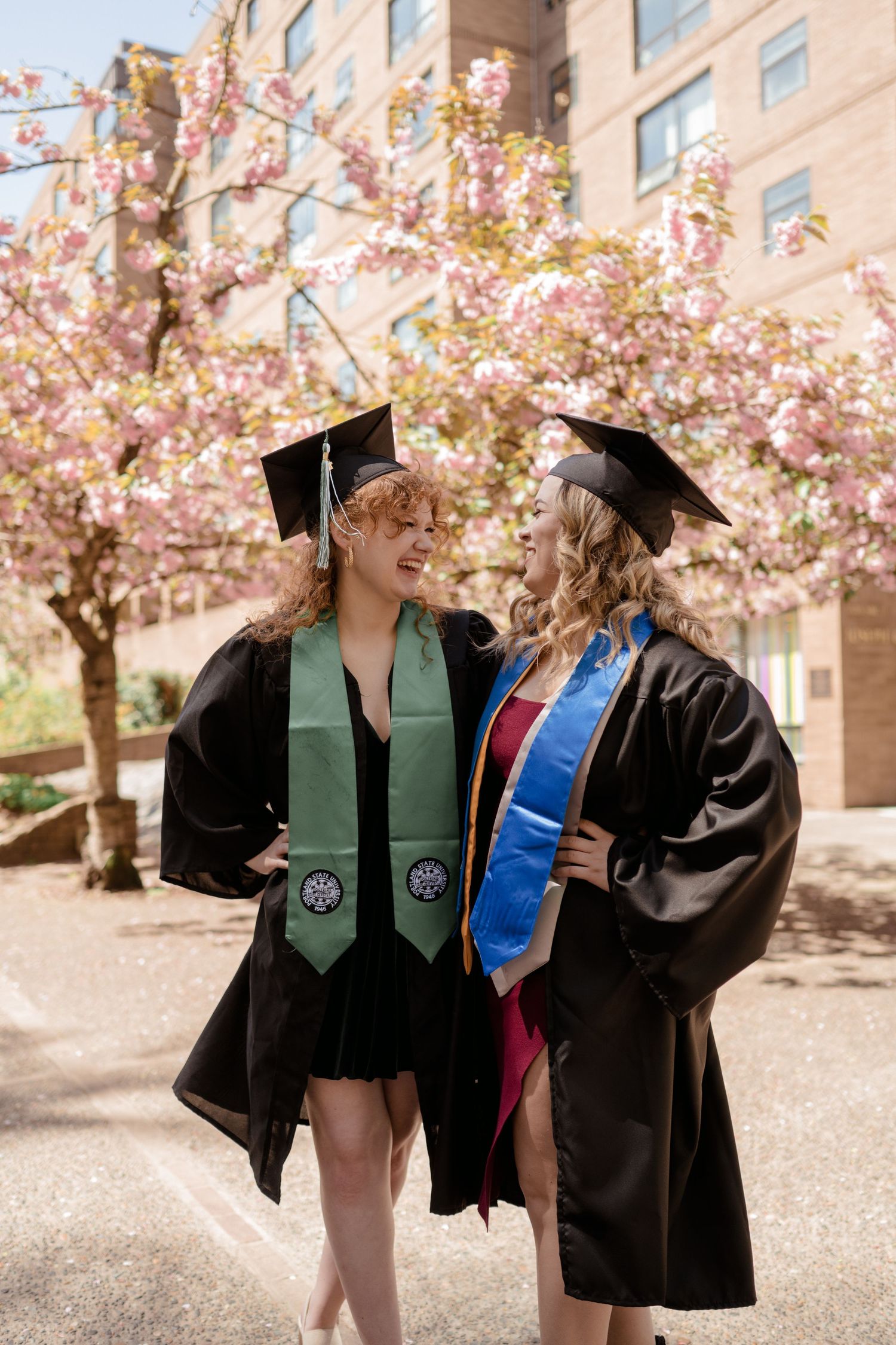 Two graduates in caps and gowns posing together under blooming cherry trees.