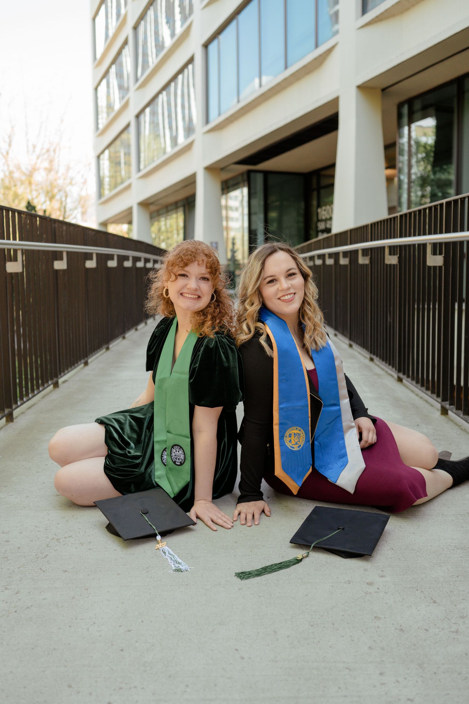 Graduates sitting on ground with caps and colorful stoles outside university building.