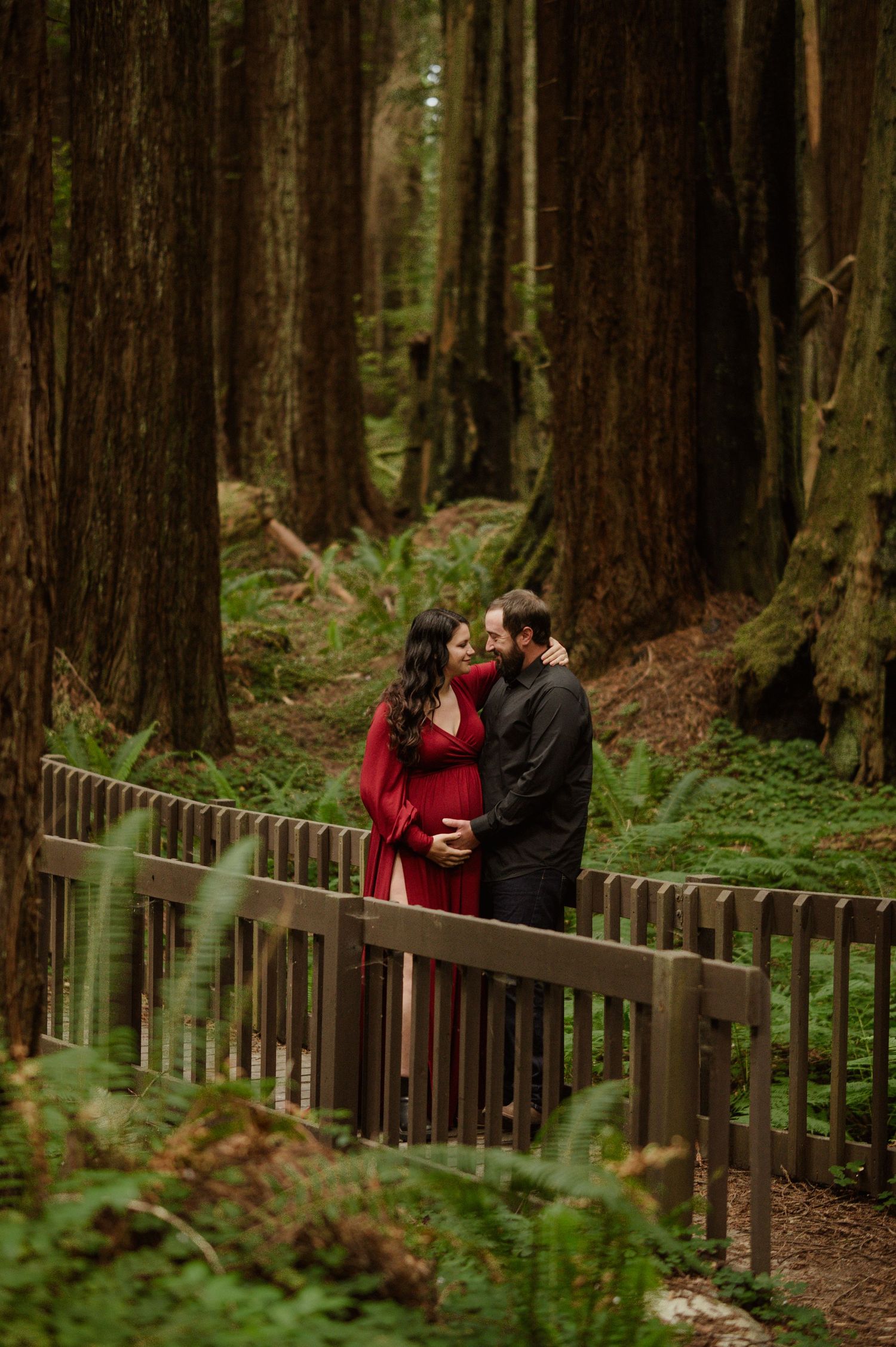 A couple shares a romantic moment on a wooden boardwalk surrounded by towering redwood trees in a misty forest.