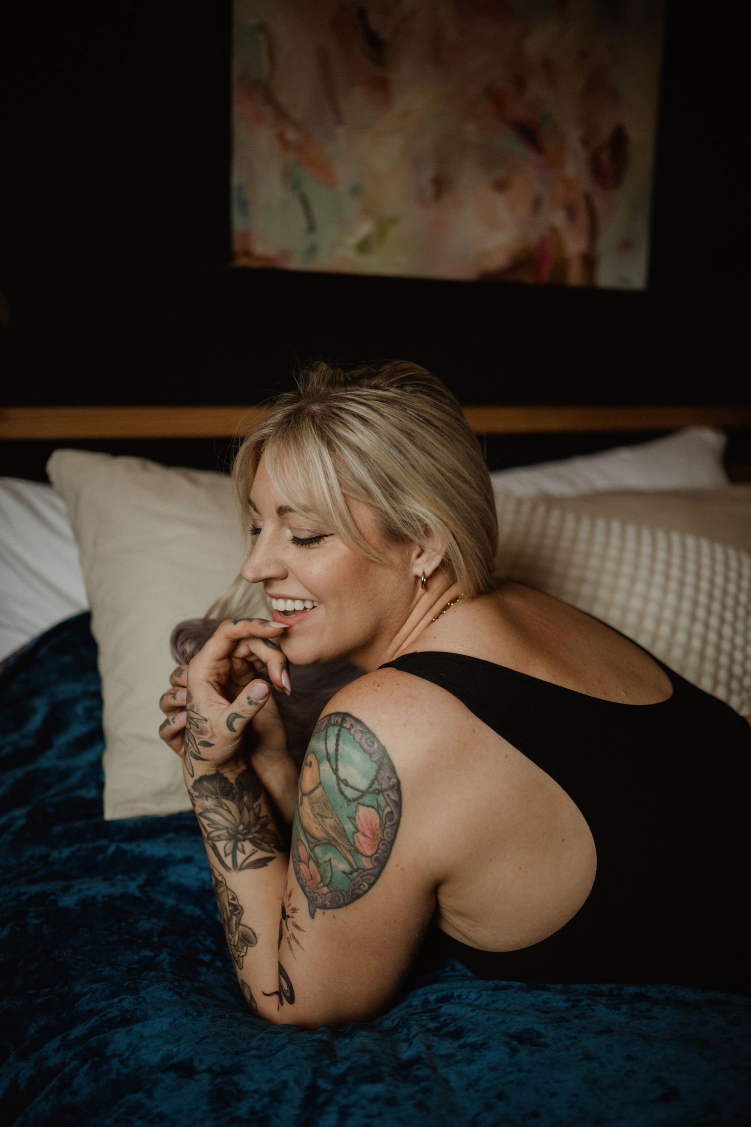 Candid portrait of blonde woman with colorful arm tattoos relaxing on blue bedding.