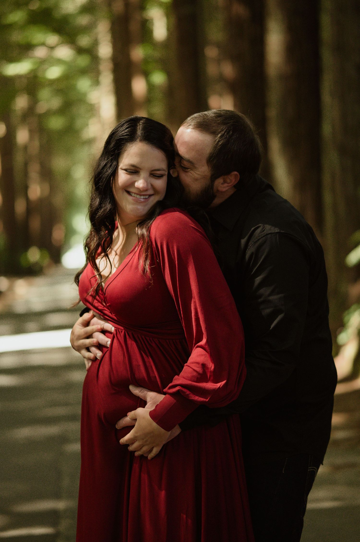 A loving couple embraces in a forest setting as sunlight filters through the trees, the woman wearing a flowing red dress.