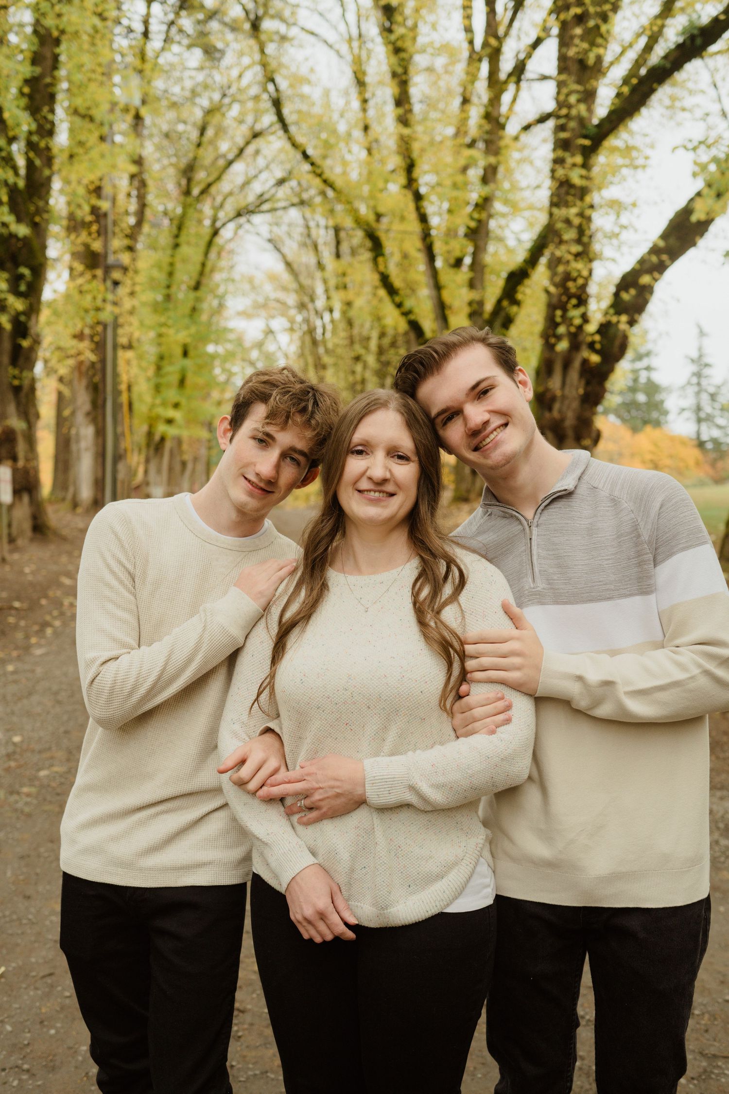 Three people in matching cream sweaters pose for a casual portrait among autumn trees.
