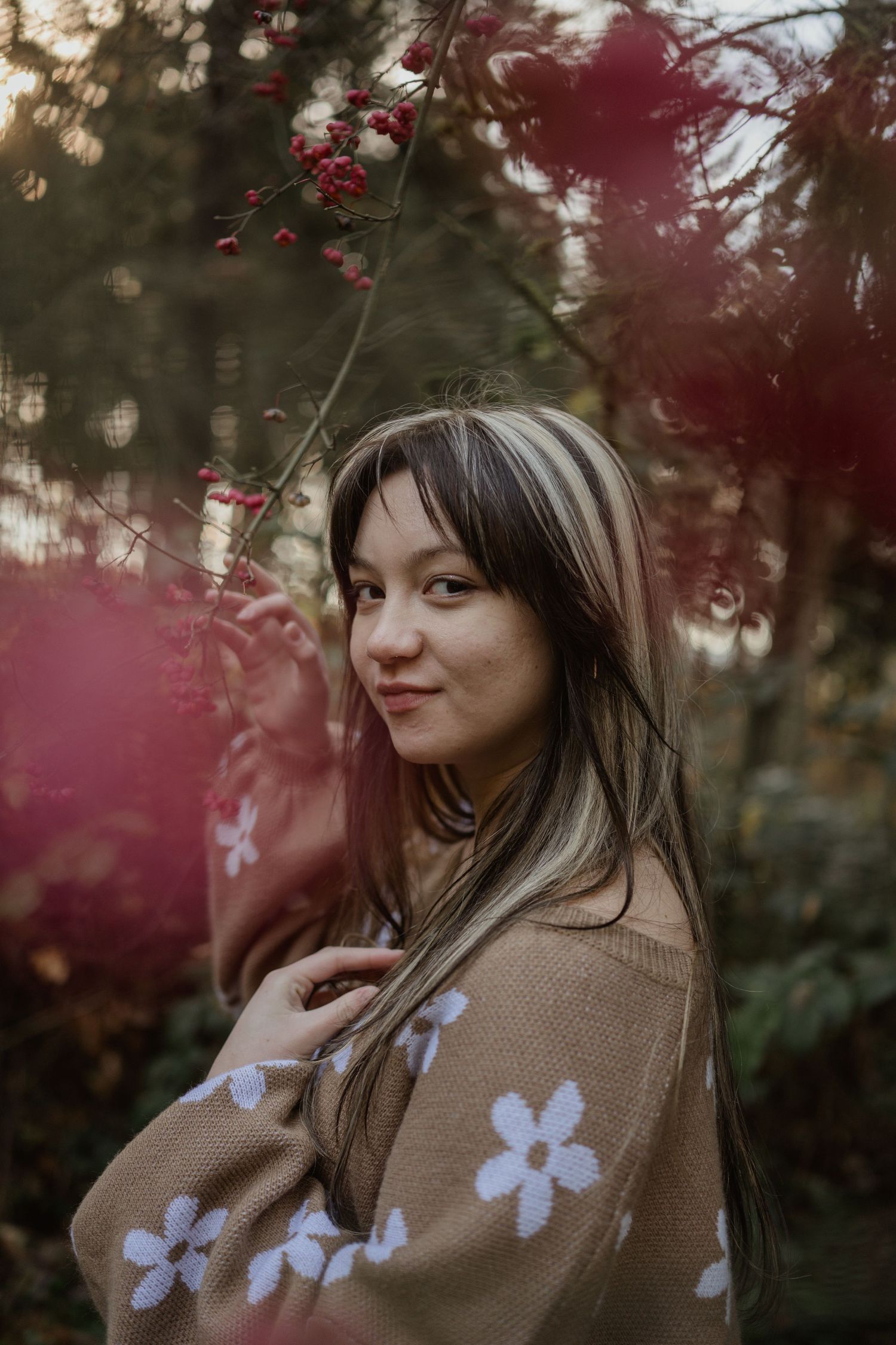 A person in a floral sweater smiles while standing among cherry blossoms in a garden.