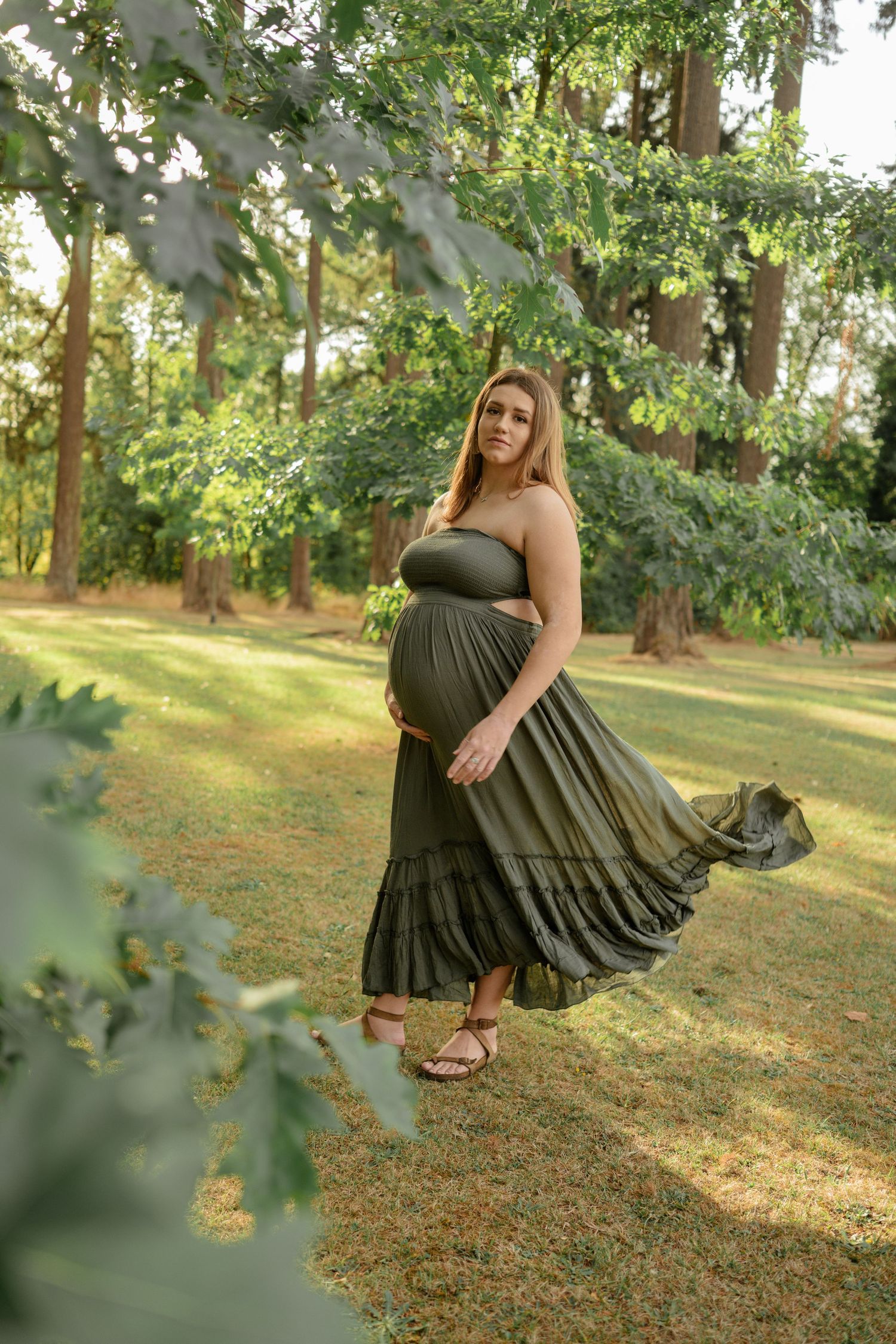 An expectant woman in an olive green maxi dress twirls in a sunlit garden setting during a maternity photoshoot.