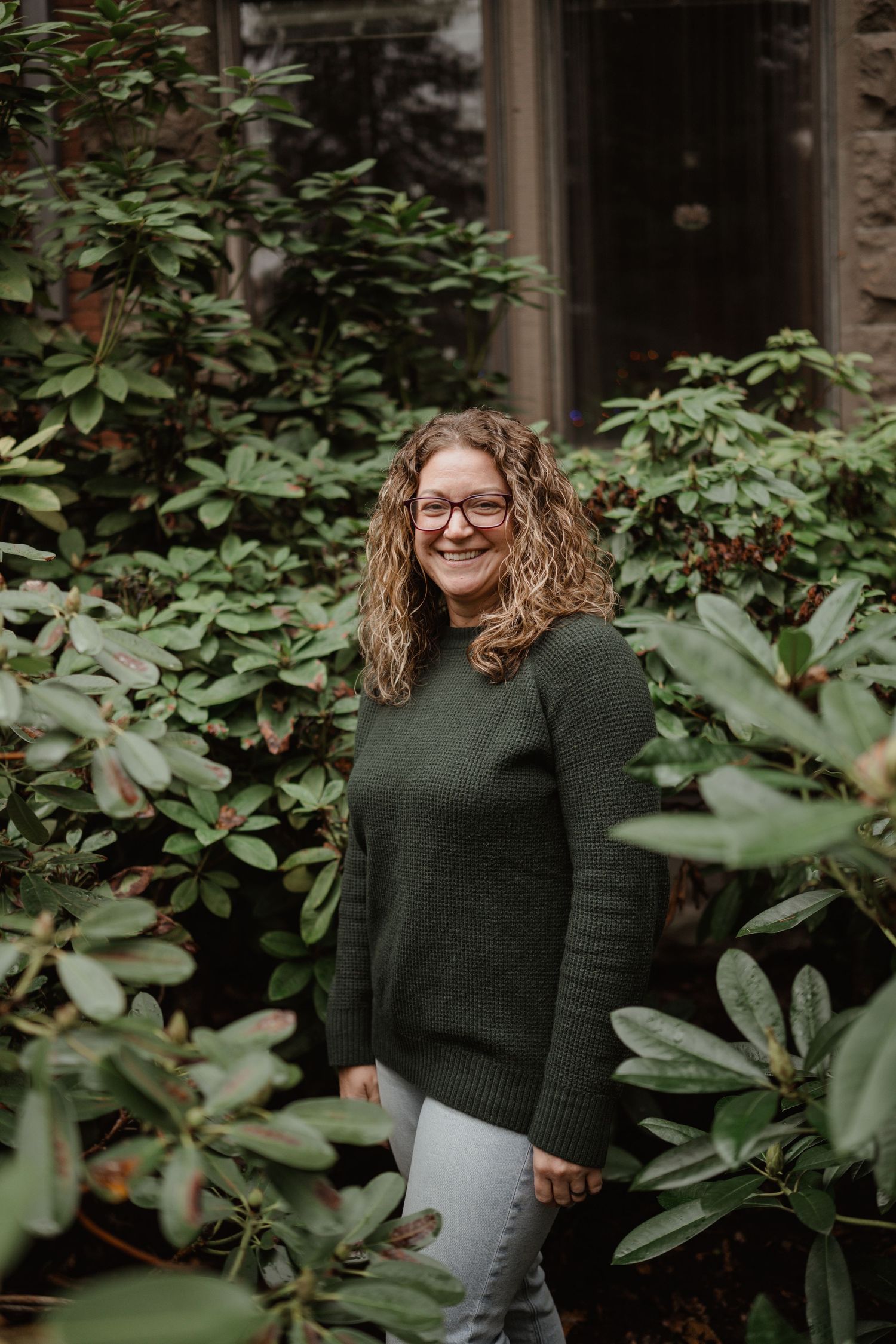 Woman in green sweater smiles while standing among lush garden foliage.