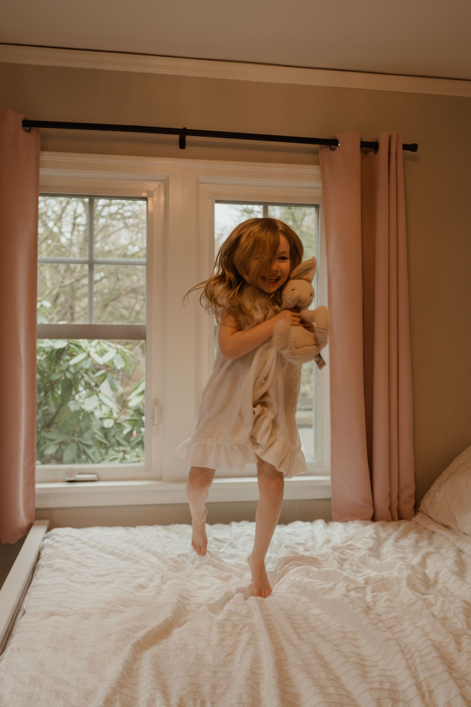 Young child in white nightgown jumping on bed near window with pink curtains.