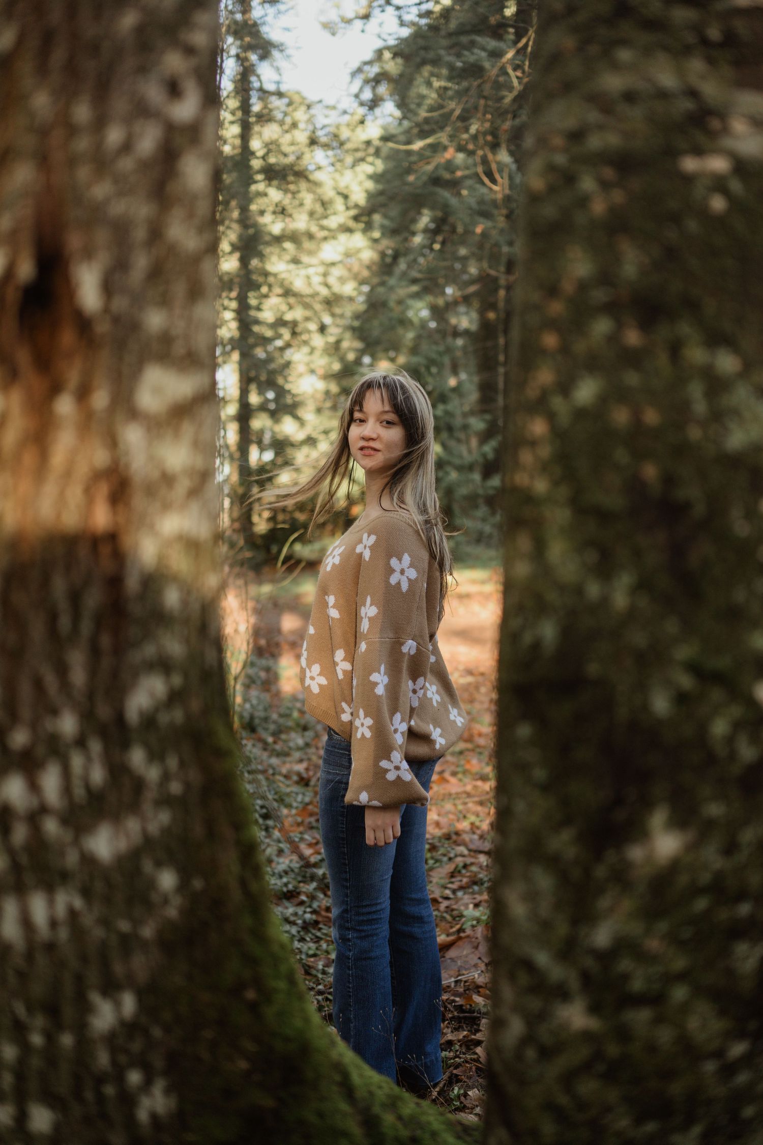 A person wearing a brown floral sweater poses between trees in autumn sunlight.