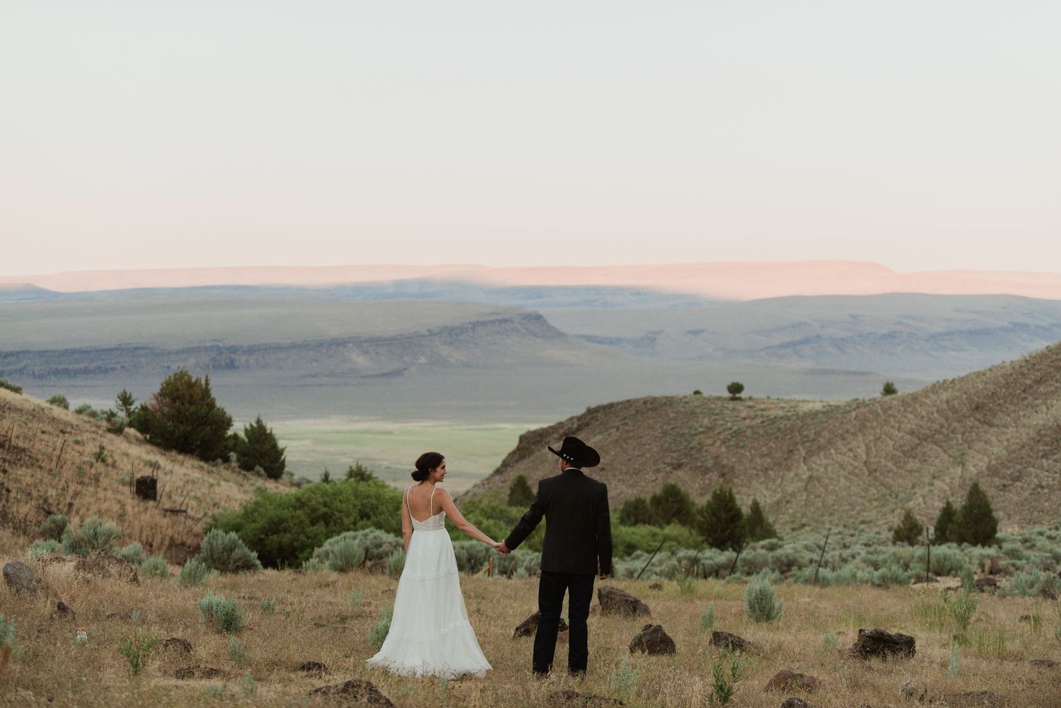 Newlywed couple stands on hilltop overlooking scenic mountain valley at sunset.