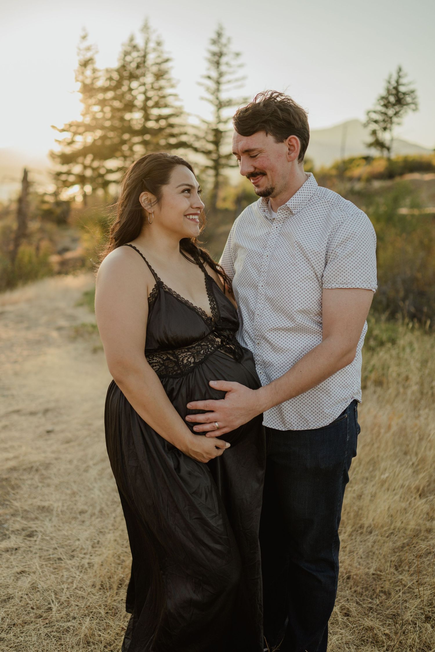 A couple poses together in a natural outdoor setting at sunset with evergreen trees in the background.