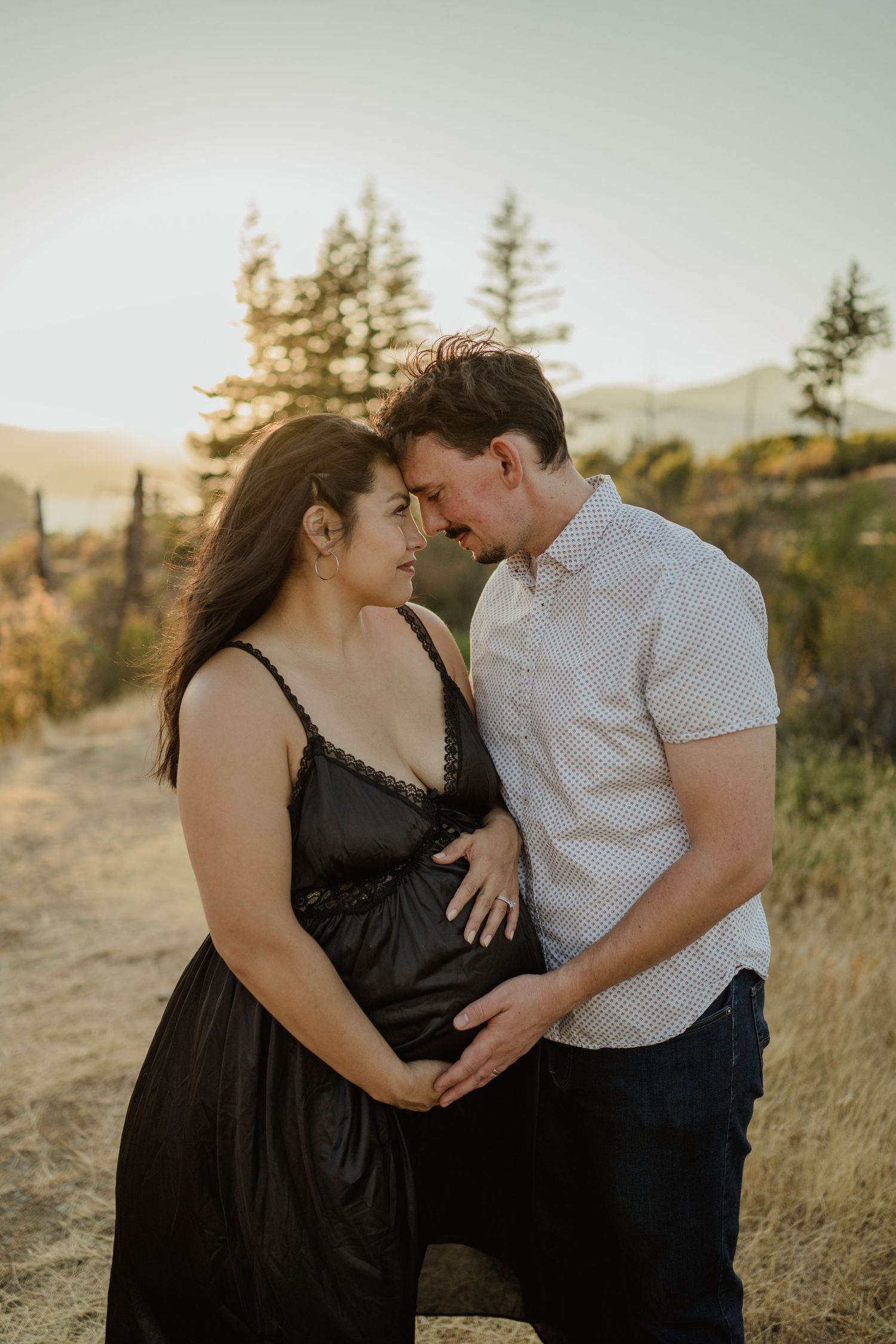 A tender moment between a couple standing on a mountain trail at sunset with evergreen trees in the background.