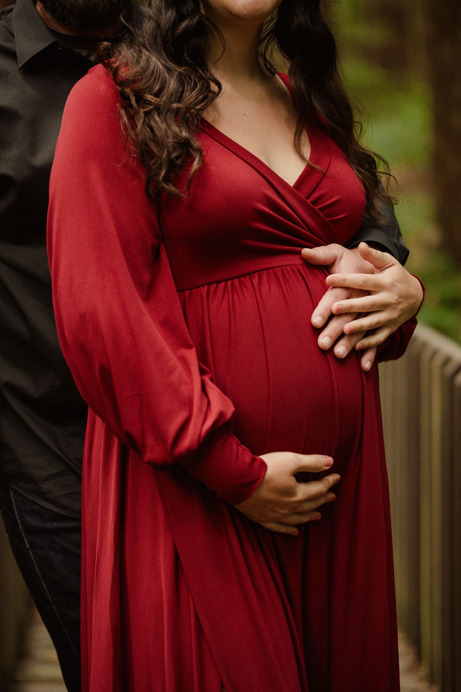 A pregnant woman in a flowing red dress holds her belly while posing for a maternity photo outdoors.
