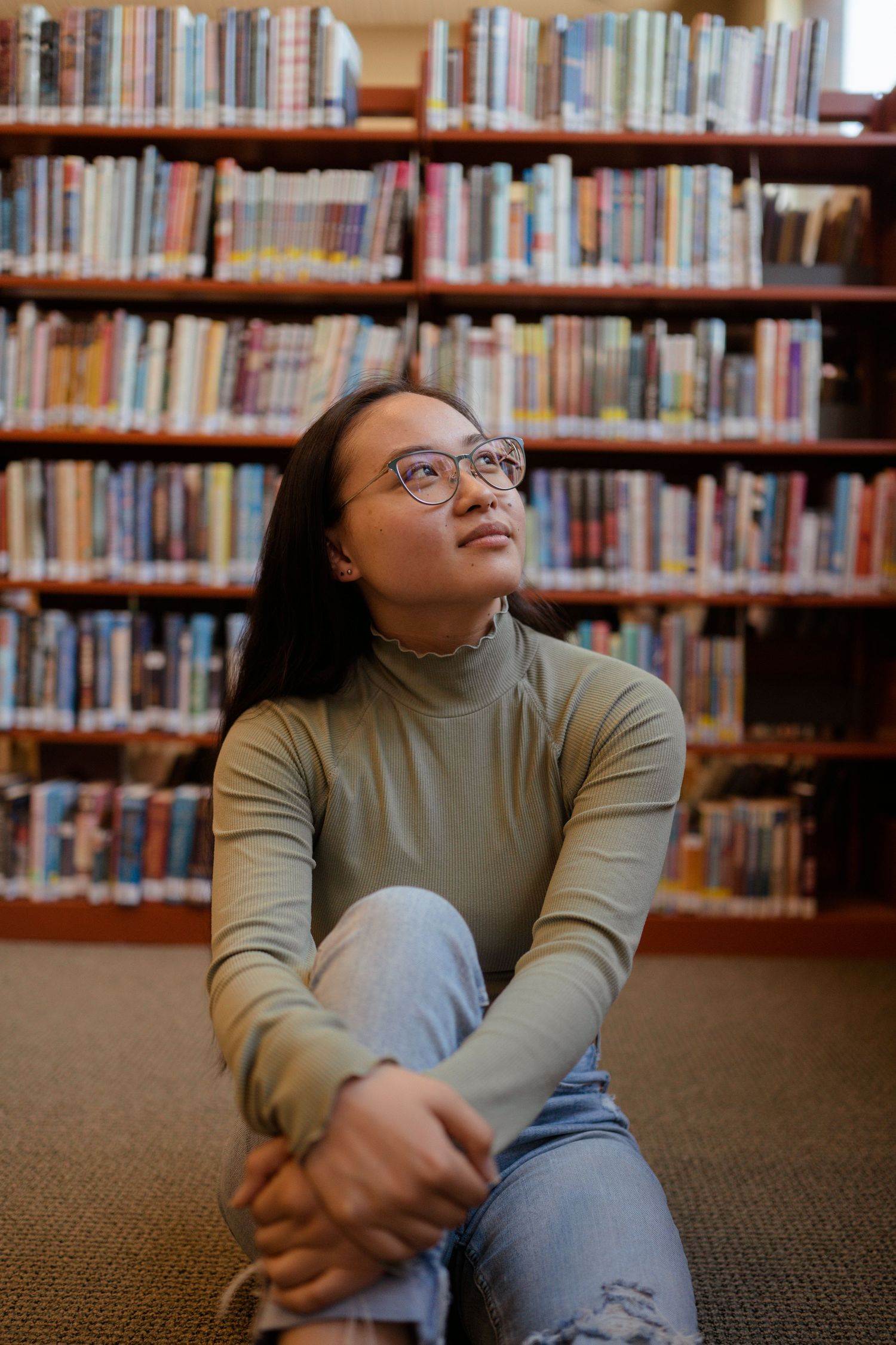 Student sitting on library floor surrounded by bookshelves wearing turtleneck and glasses looking upward thoughtfully.