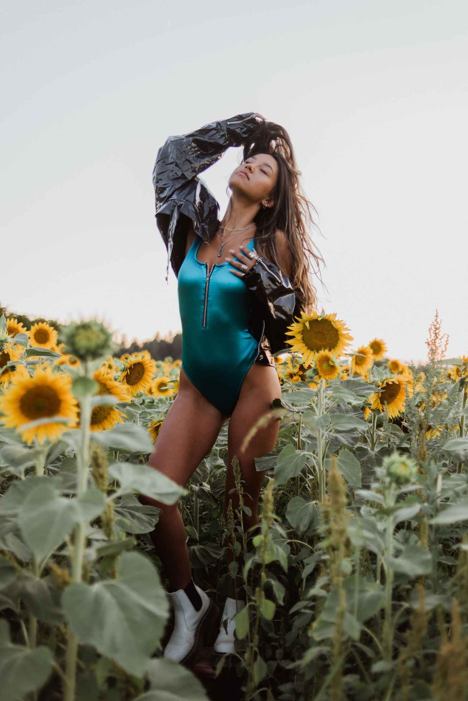 Fashion model in turquoise bodysuit and leather jacket poses dramatically in blooming sunflower field.
