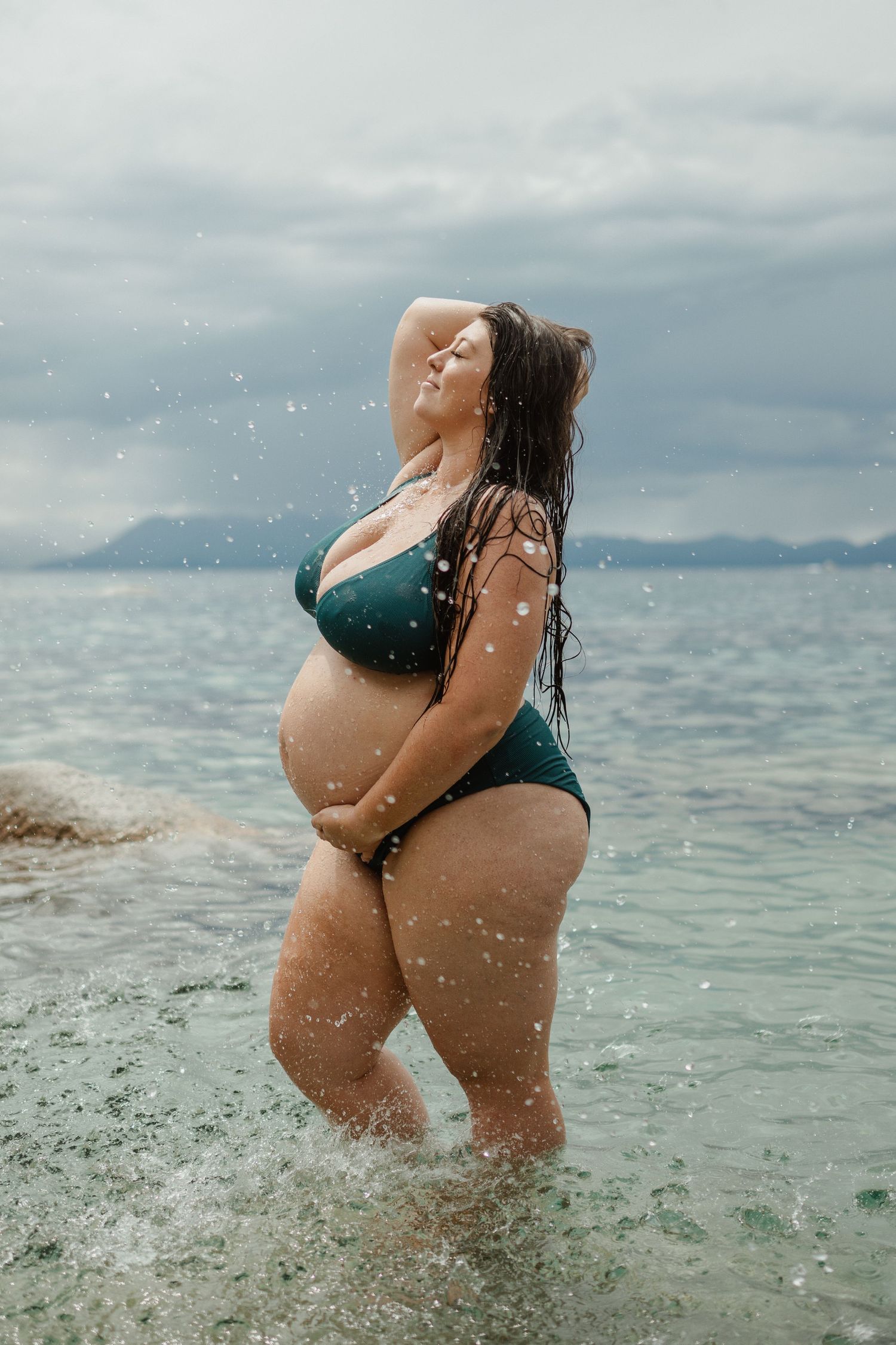 Plus size woman in emerald swimsuit enjoying ocean waves on a cloudy day at the beach.