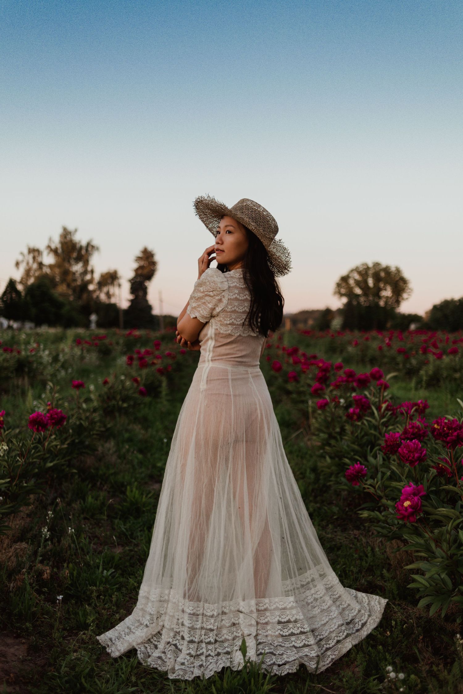 A woman in a vintage-style white lace dress and sun hat stands in a field of pink flowers at sunset.