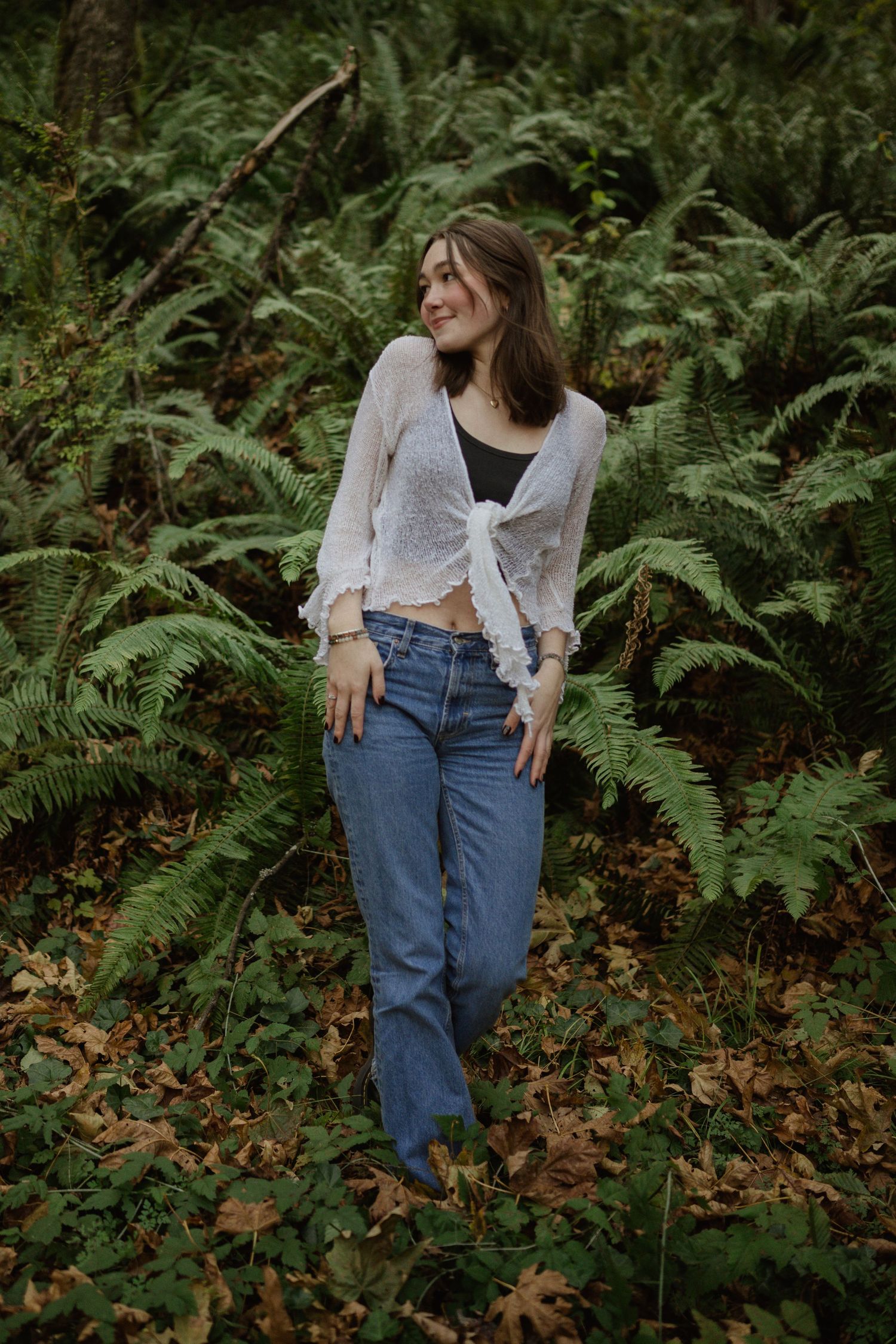 Fashion model poses in casual denim and white cardigan among lush green ferns in forest setting.