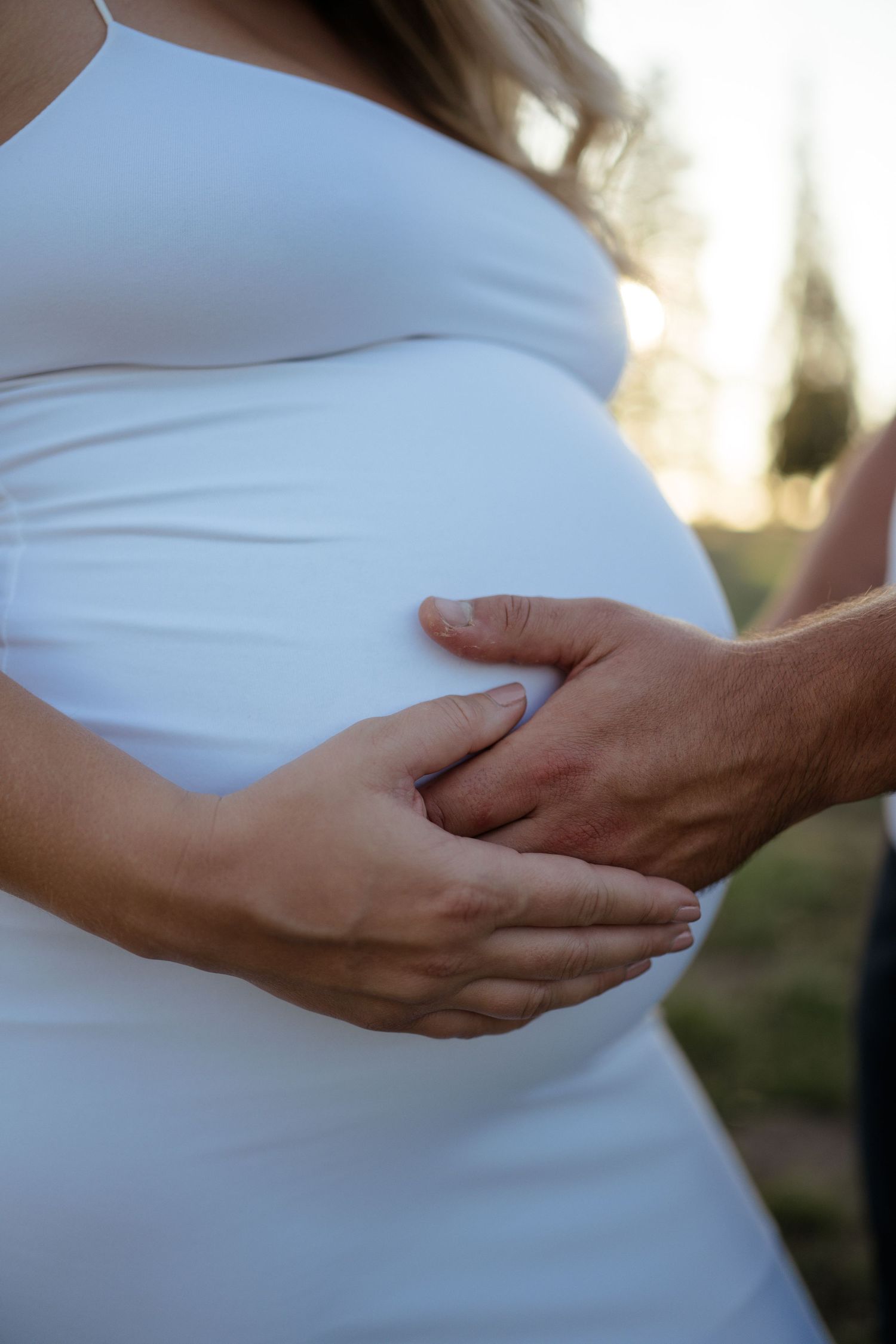 Close up of hands tenderly holding pregnant belly in white maternity dress during outdoor photo session.