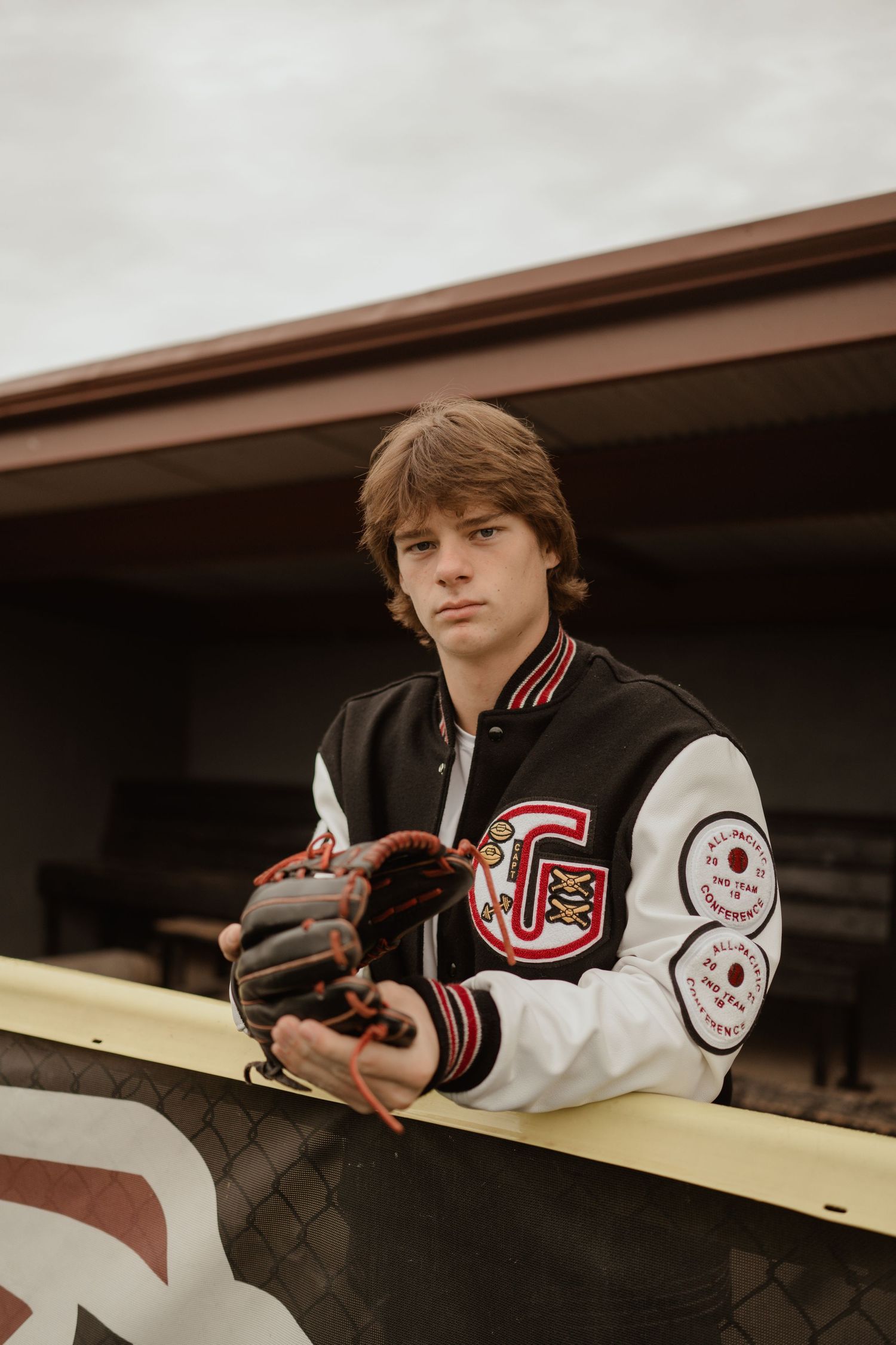 Baseball player in letterman jacket leans on dugout railing holding a baseball glove.