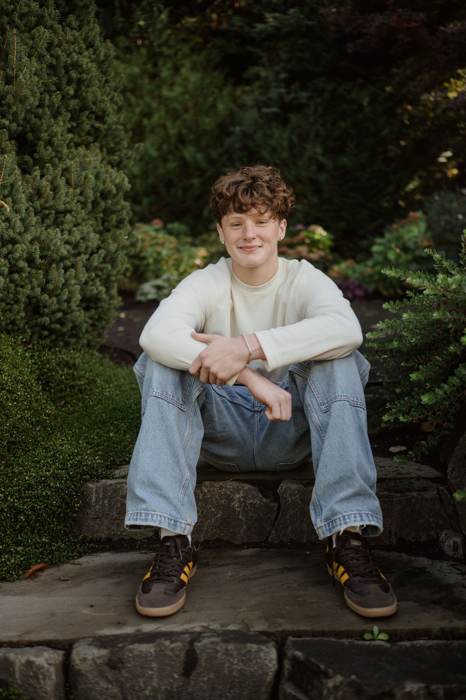 A casual portrait of someone sitting on stone steps in a garden wearing jeans and a cream sweater.