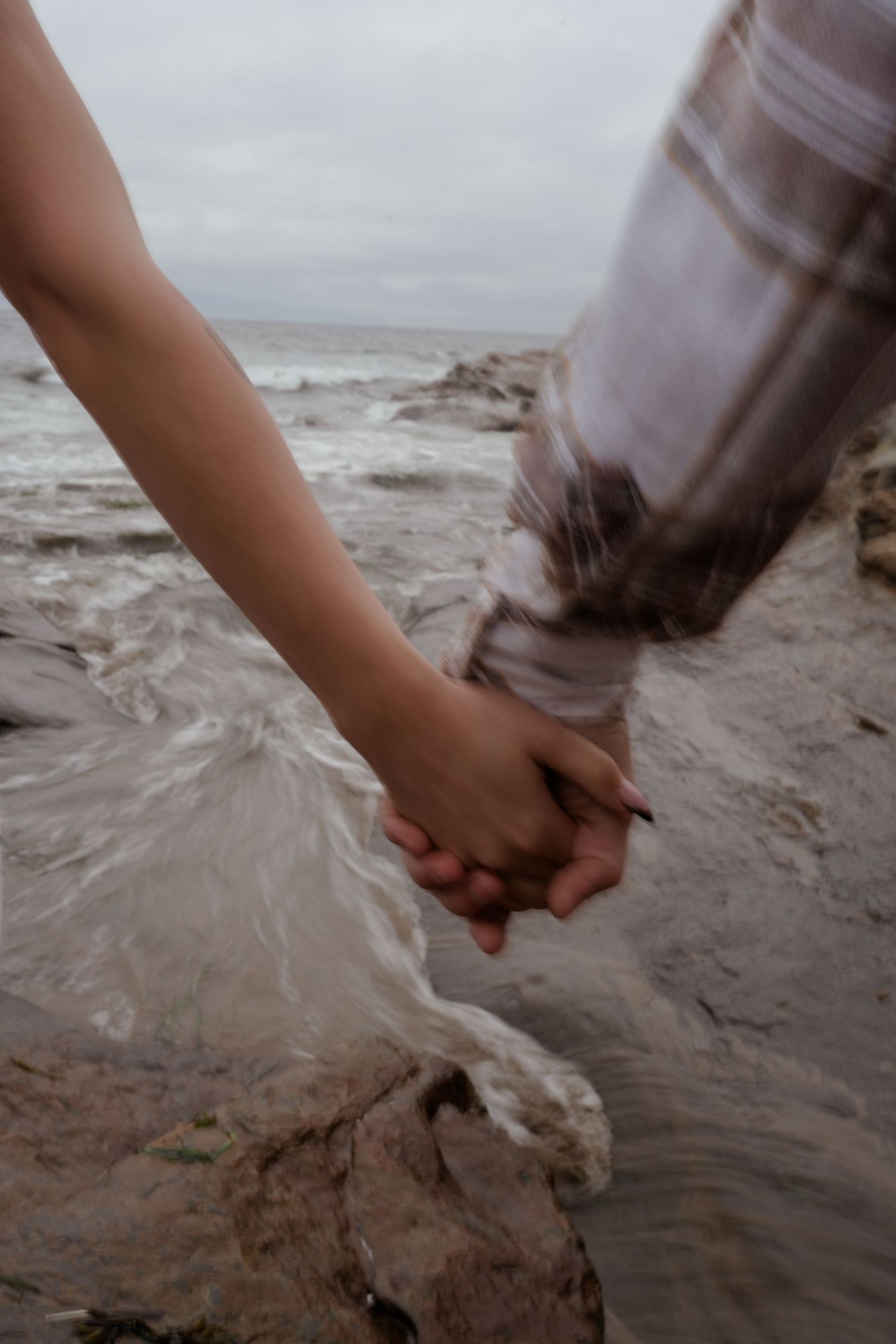 Close-up shot of two hands clasped together on a rocky beach shoreline.