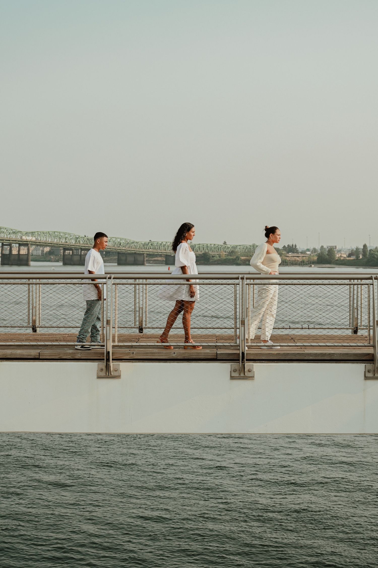 Three people in white outfits standing on waterfront boardwalk at sunset.