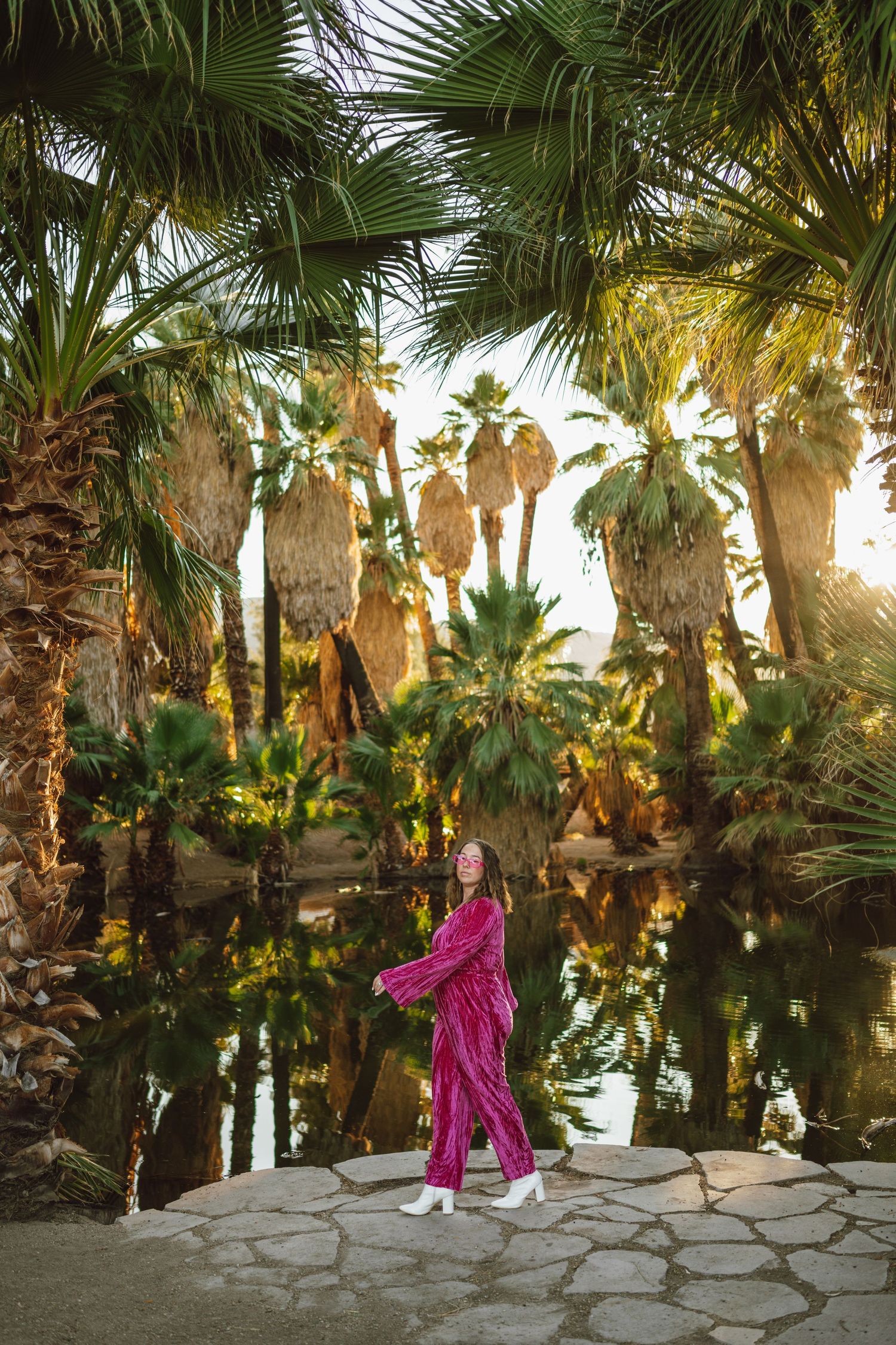 A person in pink stands among tropical palm trees beside a reflective pond.