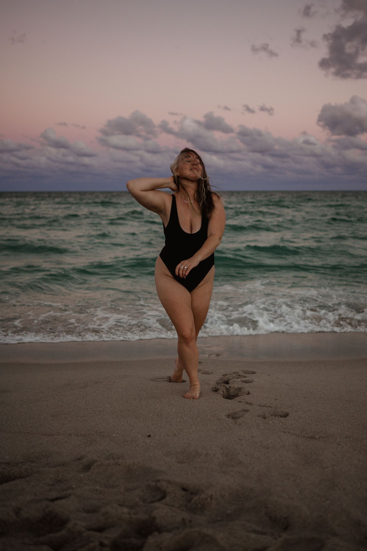 A person in a black swimsuit poses on the beach at sunset with moody clouds and turquoise ocean waves in the background.