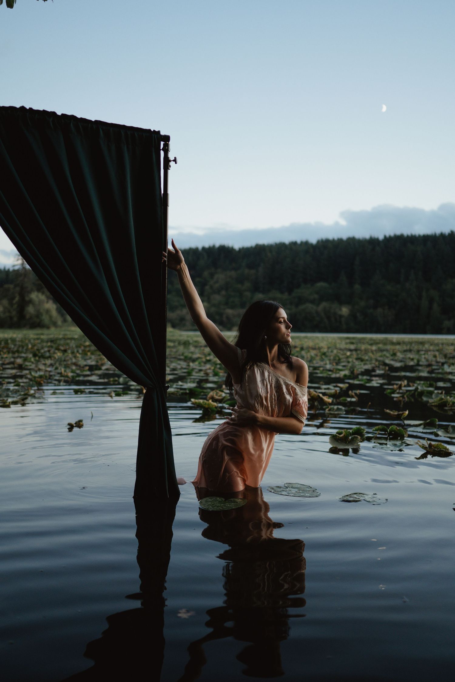 A person stands in a reflective lake with lily pads at sunset, holding a dark curtain against the mountain backdrop.