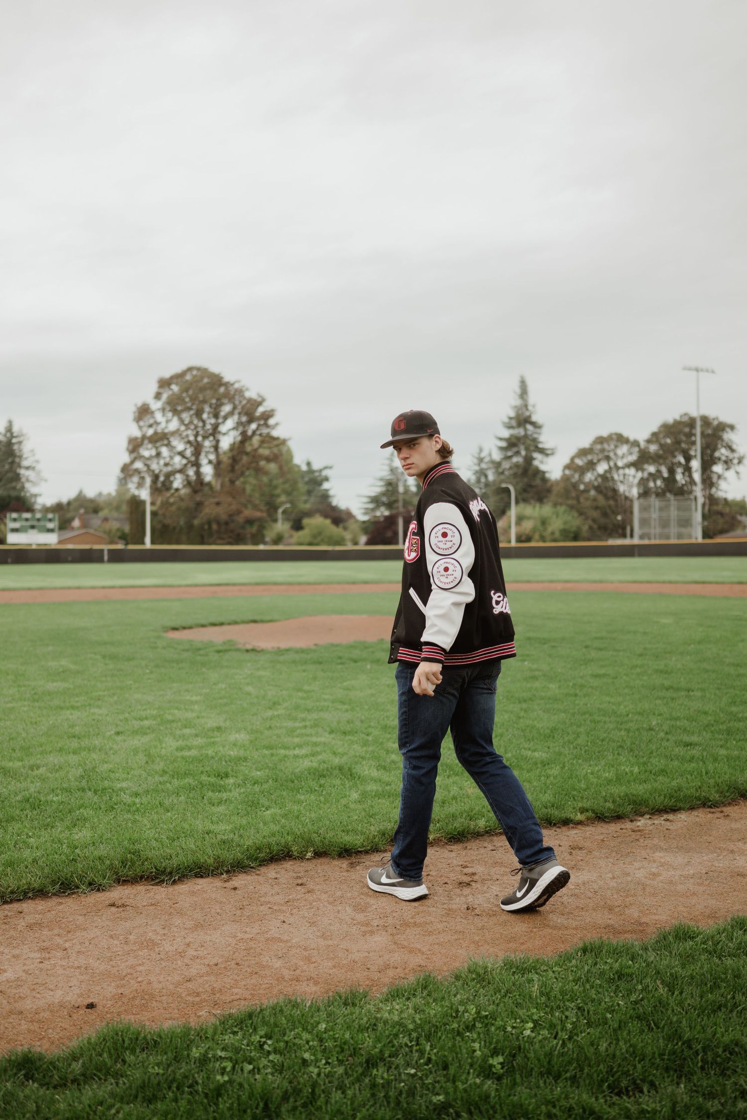 Baseball player walks along dirt path on baseball field wearing varsity jacket.