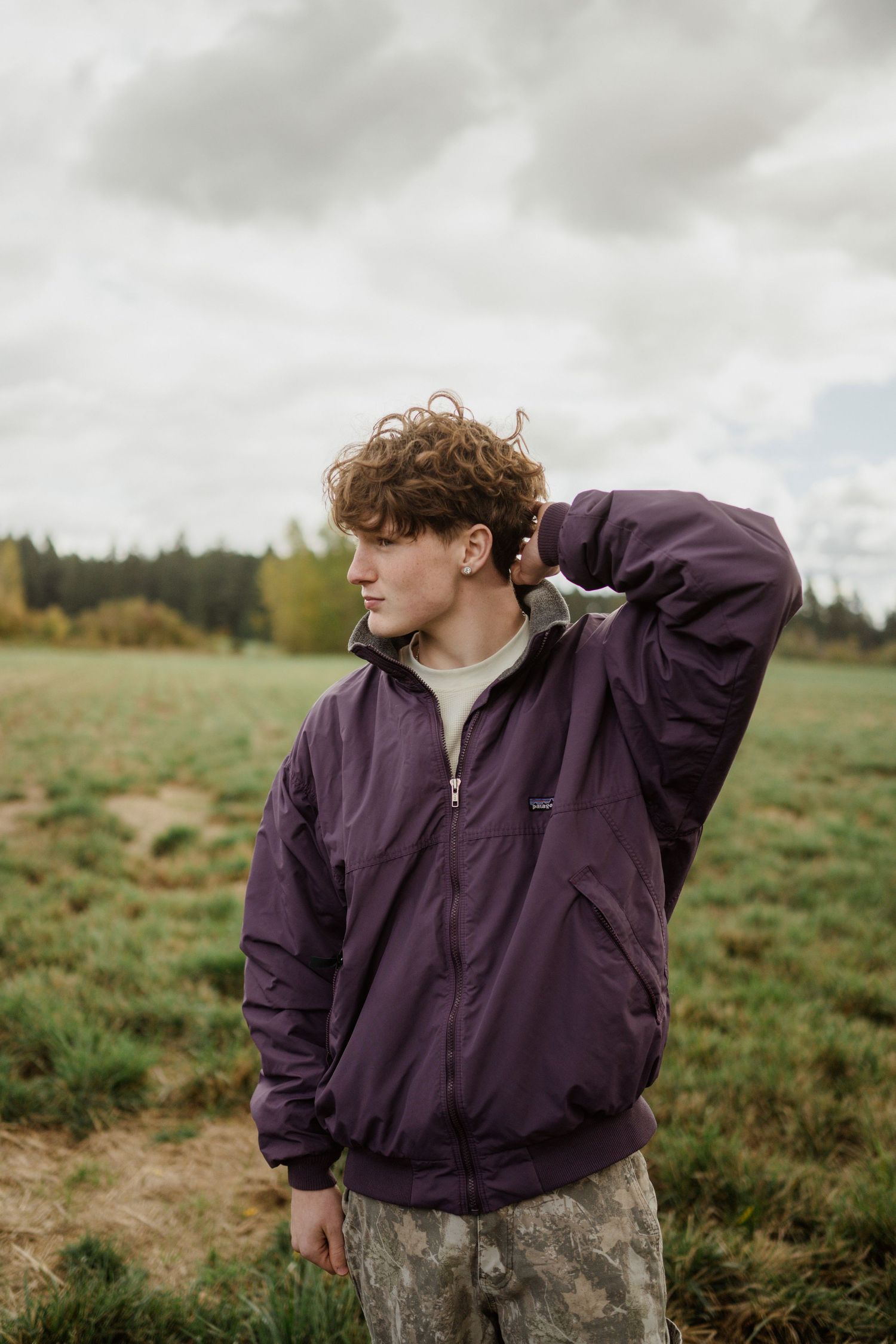 Someone wearing a purple jacket and camo pants poses in an open field with cloudy skies.