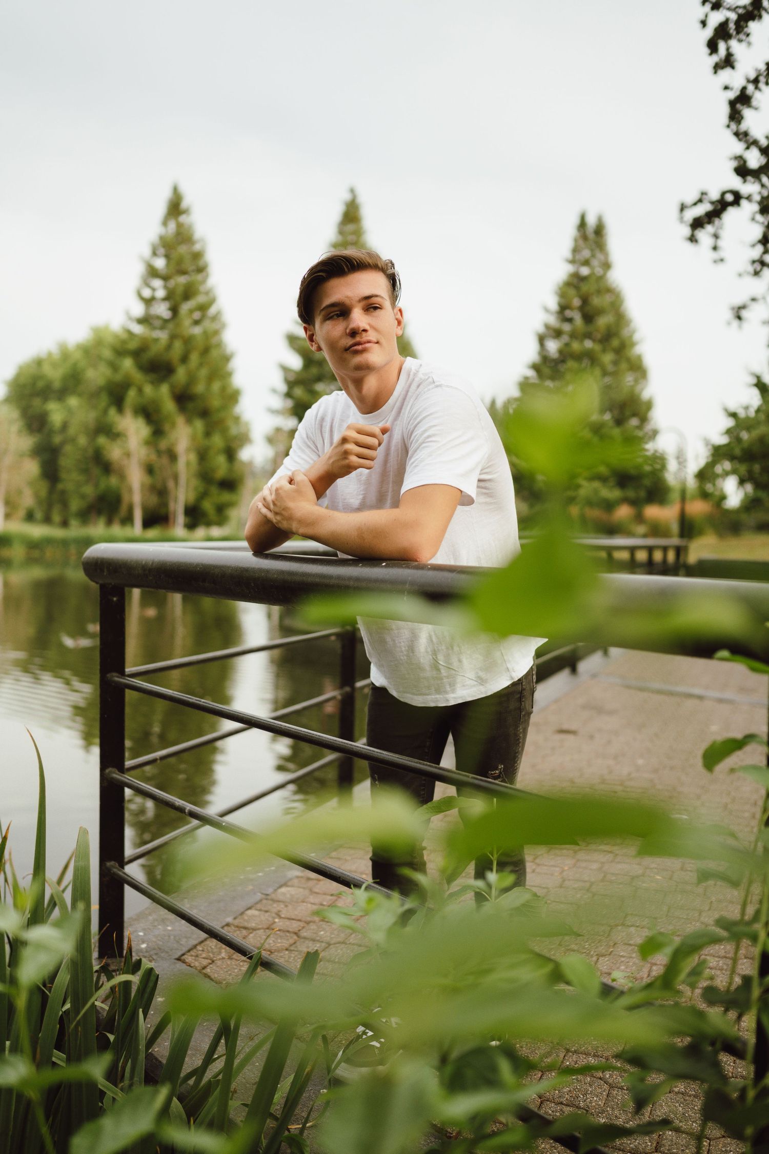 Person in white t-shirt leans casually against railing by pond with evergreen trees in background.