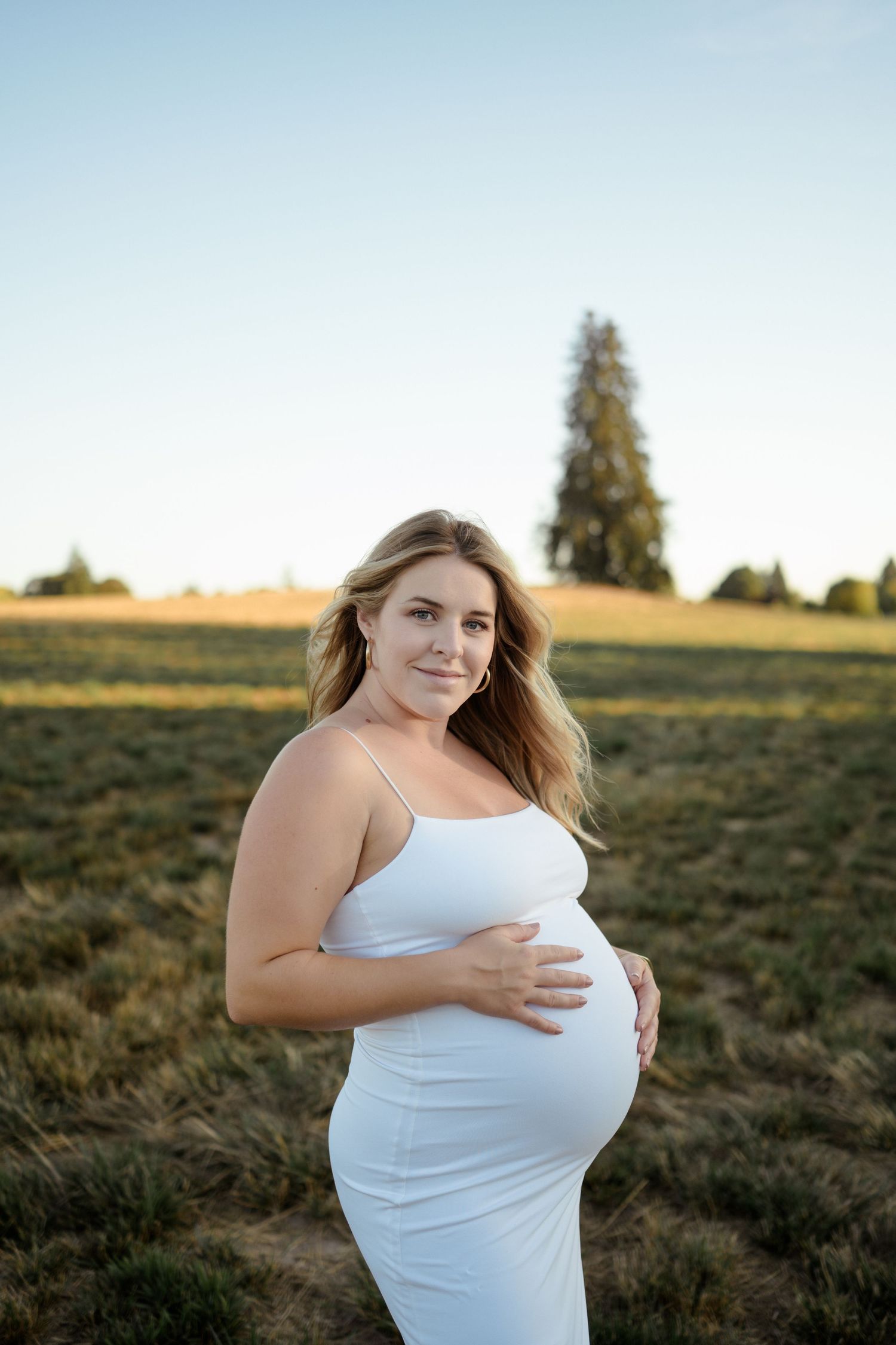 Expectant mother wearing a white dress poses in golden hour light in an outdoor field with trees in background.