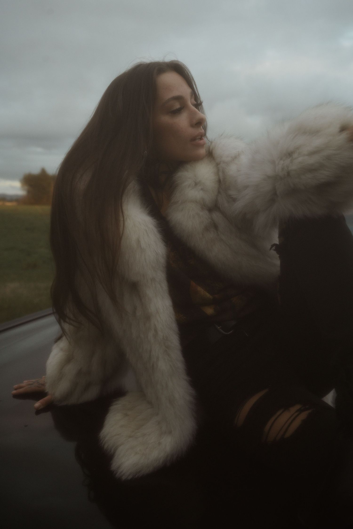 Person in a fluffy white fur coat posing outdoors on an overcast day with dramatic lighting.