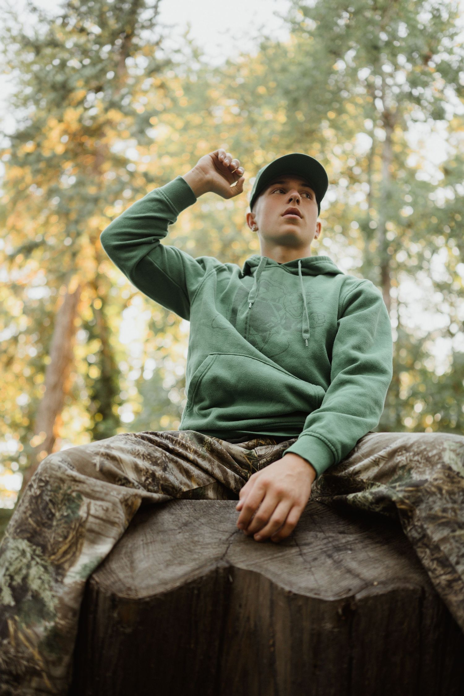 Low-angle shot of someone in a green hoodie and camo pants sitting on a large tree stump.