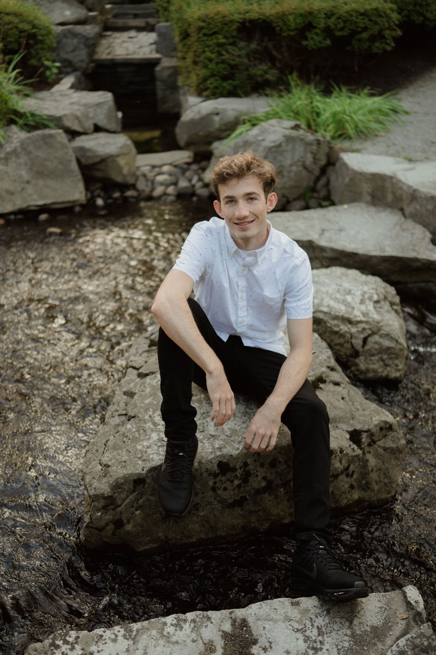 Person sitting on rocks near water feature wearing white button-up shirt and black pants.