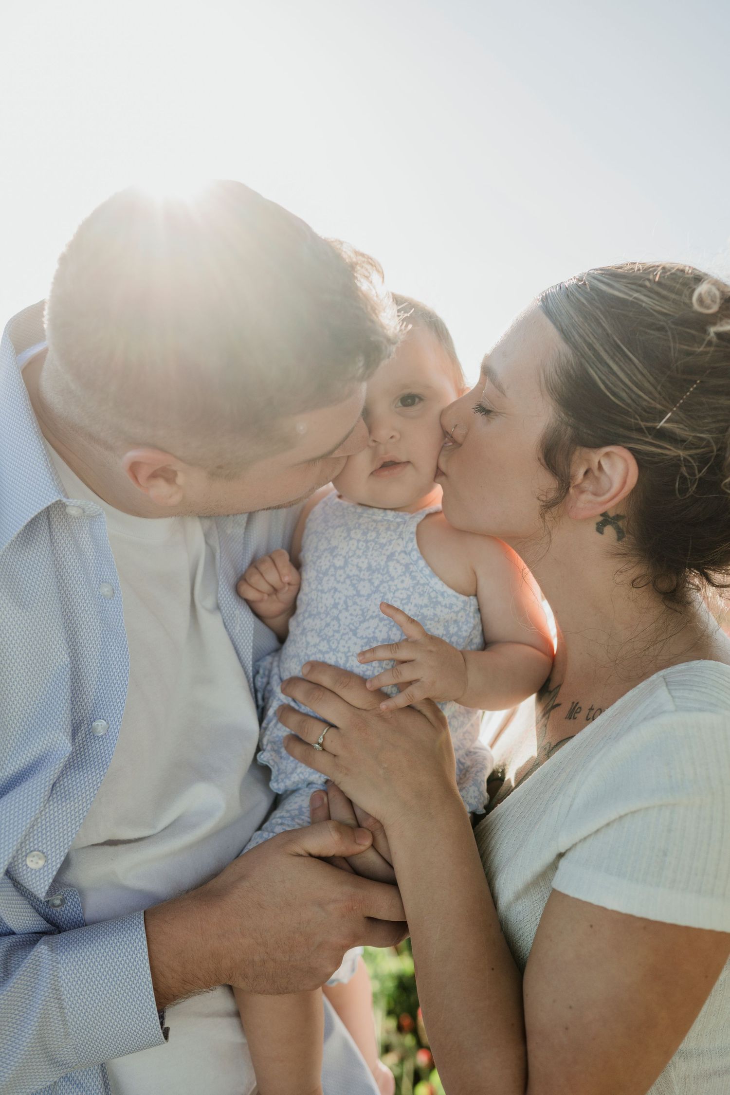 A tender family moment of parents kissing their baby in soft sunlight.