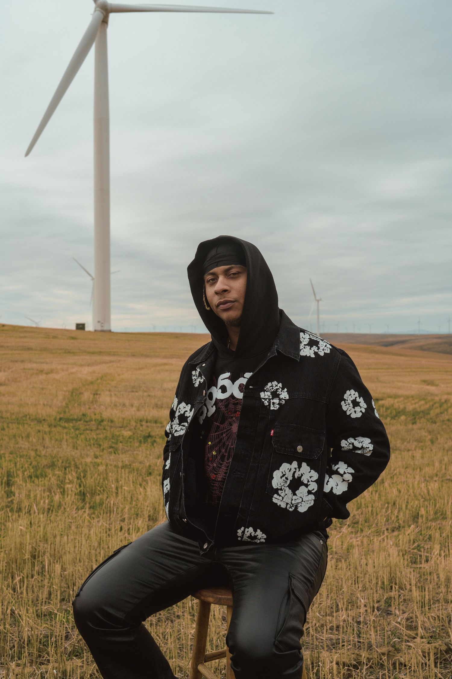 Figure in floral hoodie poses against backdrop of wind turbines in golden field.