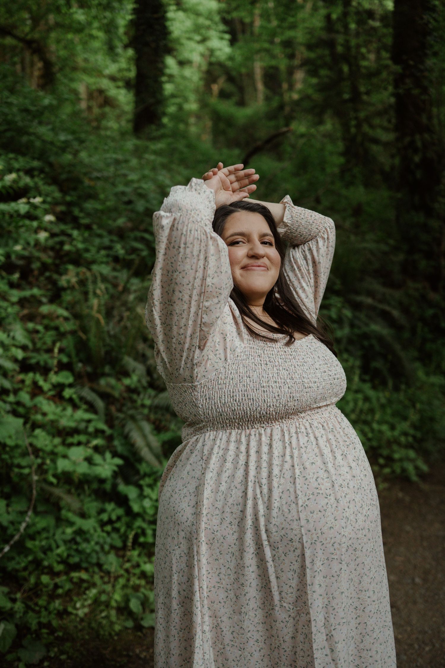 A pregnant person in a cream-colored dress strikes a joyful pose against a lush green forest background.