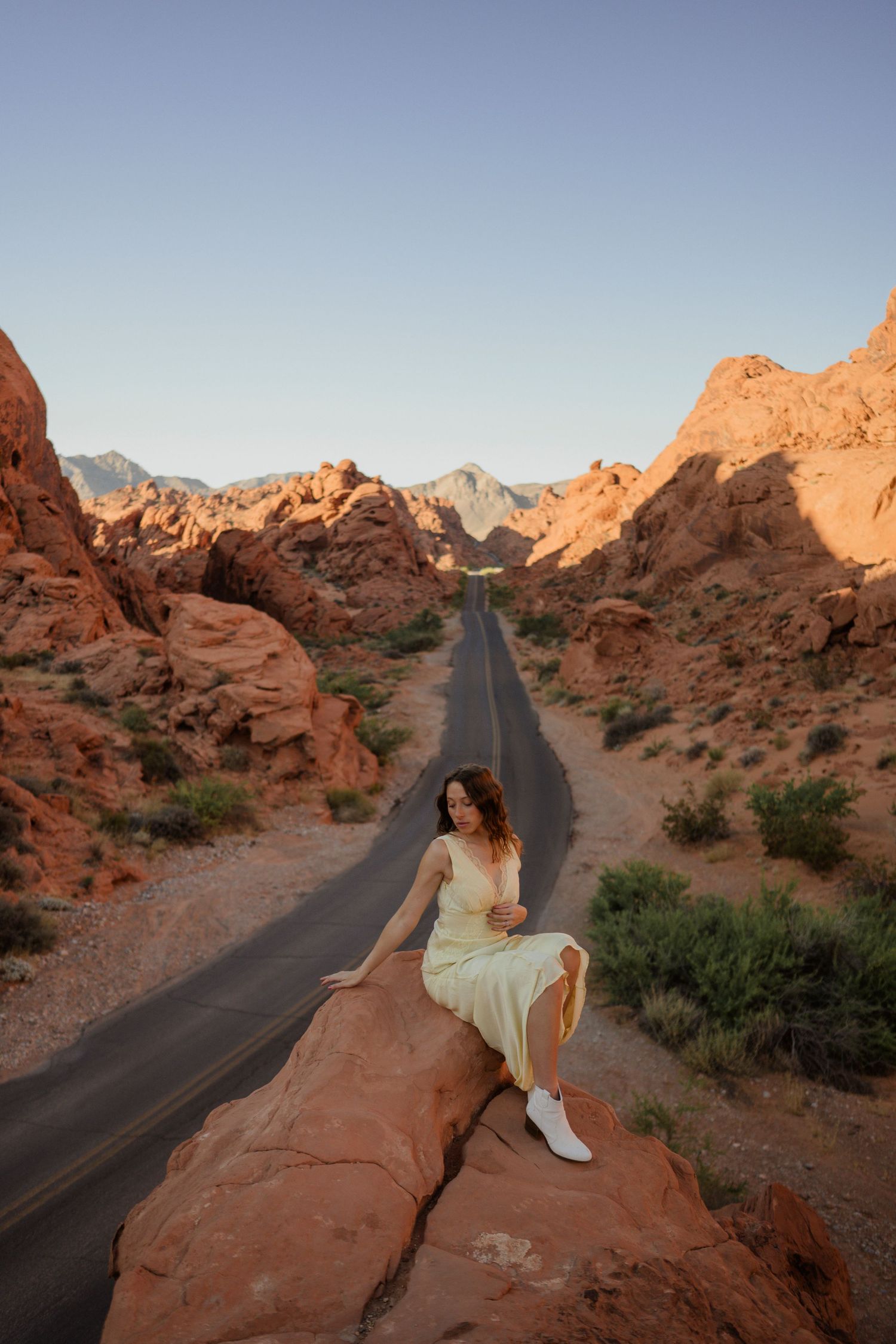 Figure in white dress sits on red rock formation overlooking desert road at sunset.