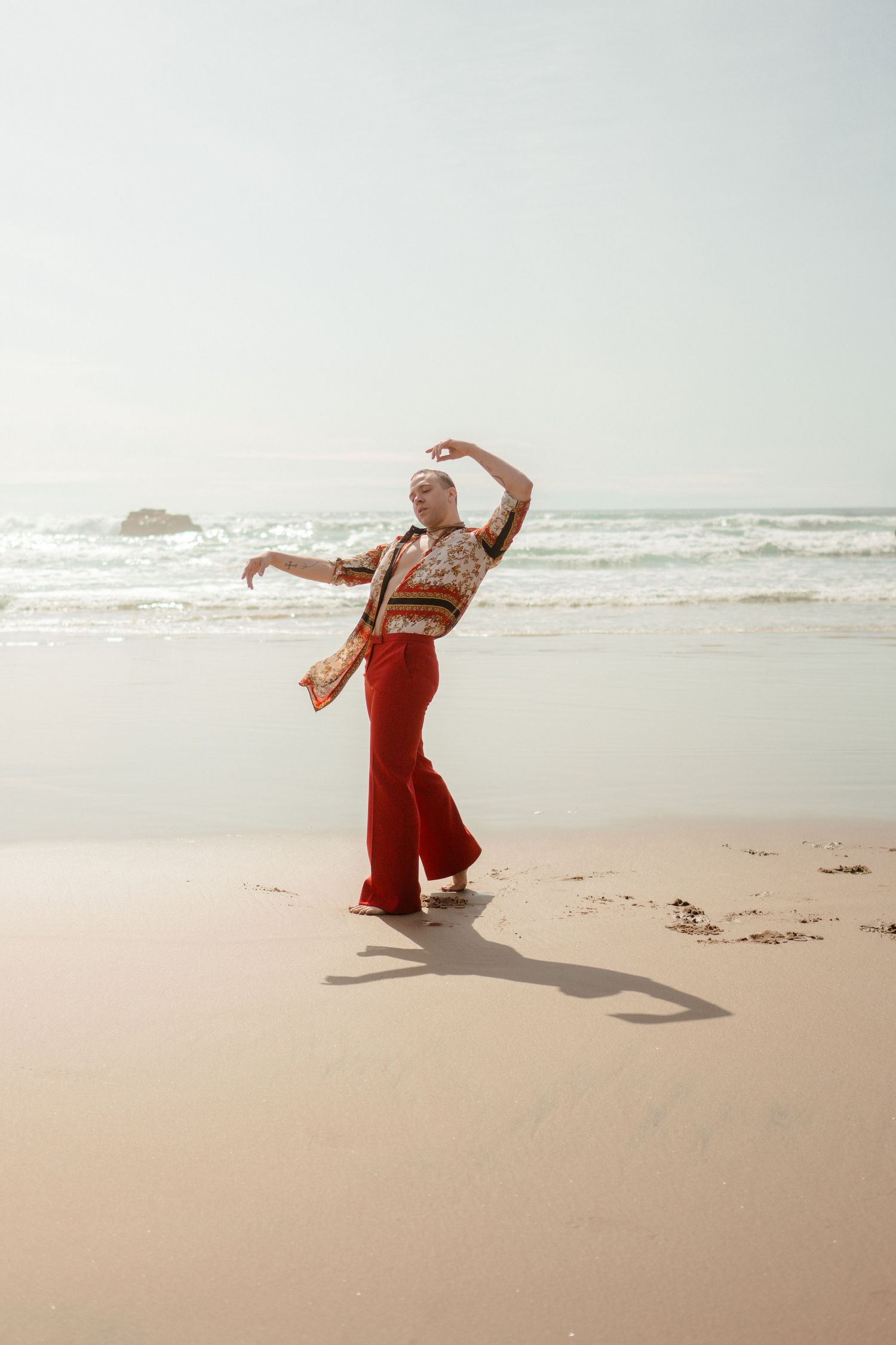 Person dancing joyfully on beach wearing red pants and striped top.
