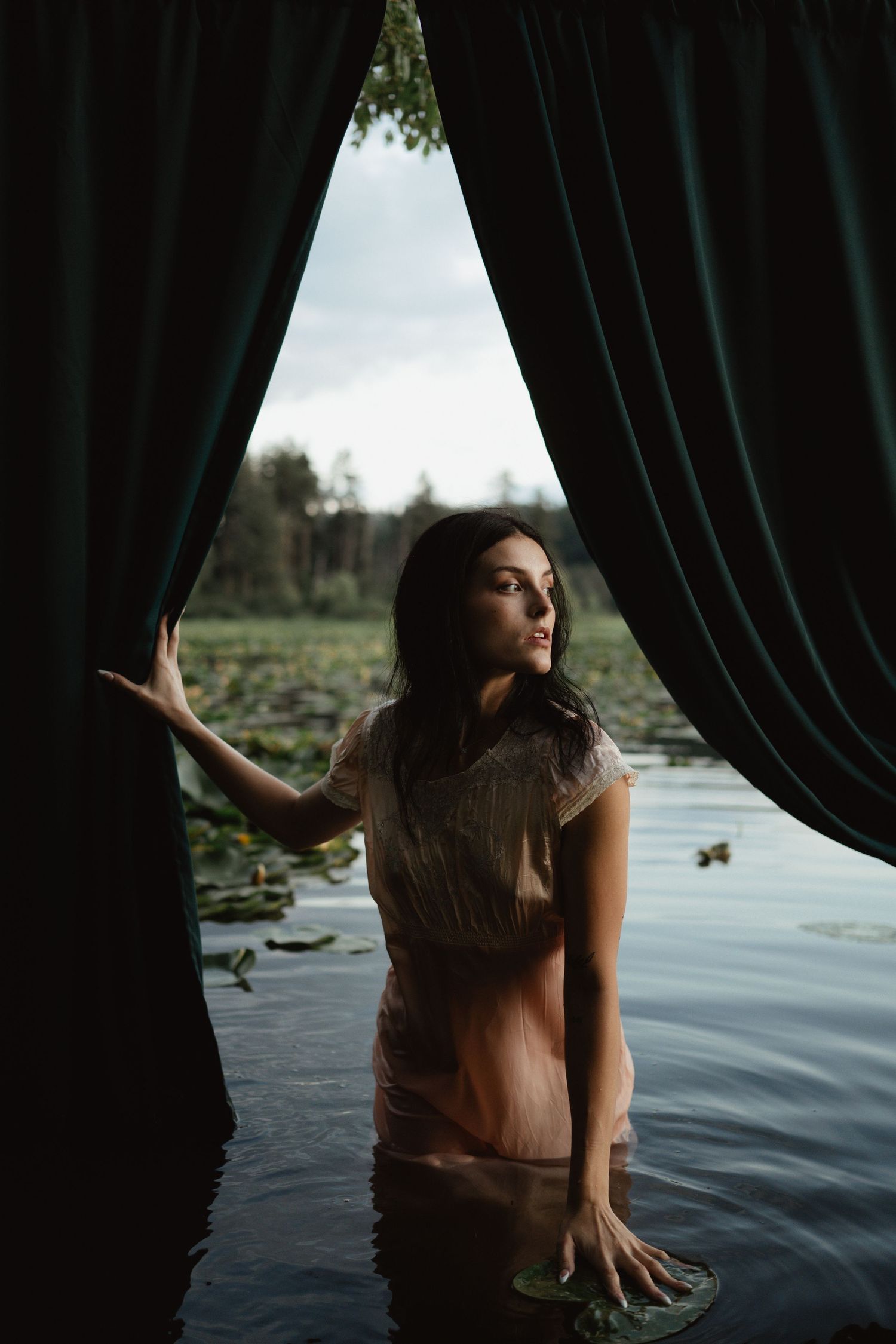 A dramatic portrait of someone standing in water behind dark curtains beside a lake with lily pads.
