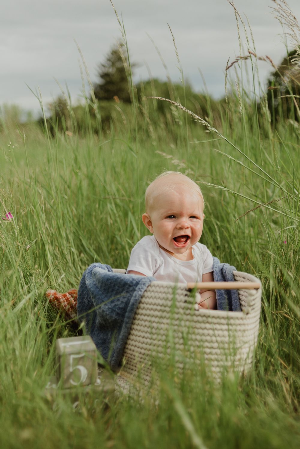 Happy baby sitting in woven basket surrounded by tall grassy field.