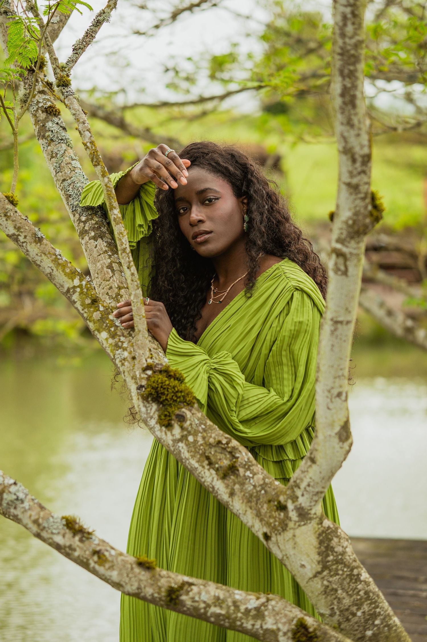 Ethereal portrait in a lime green flowing dress posed gracefully among mossy tree branches near a peaceful pond.