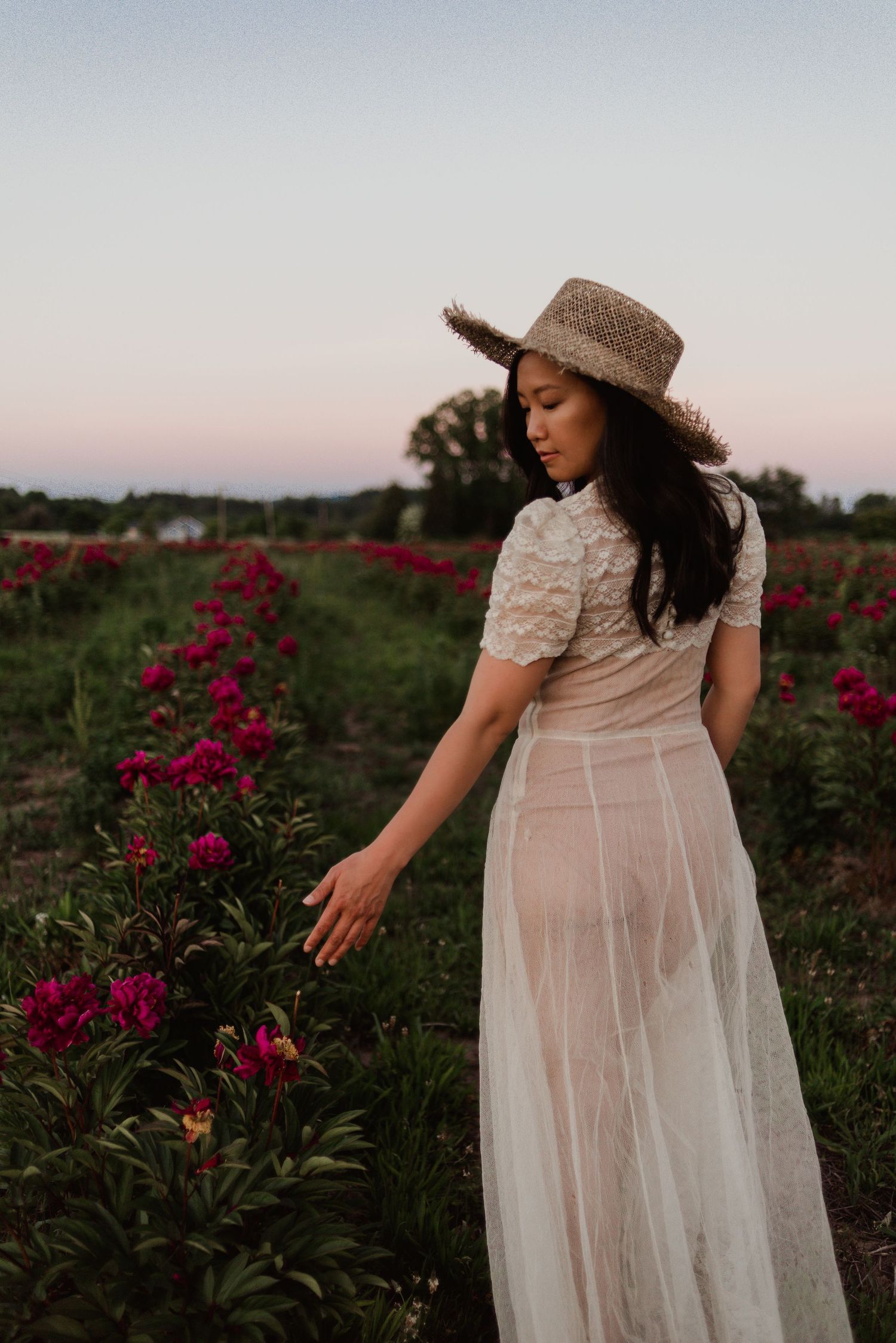 A person in a flowing white lace dress and straw hat reaches towards red flowers in a garden at sunset.
