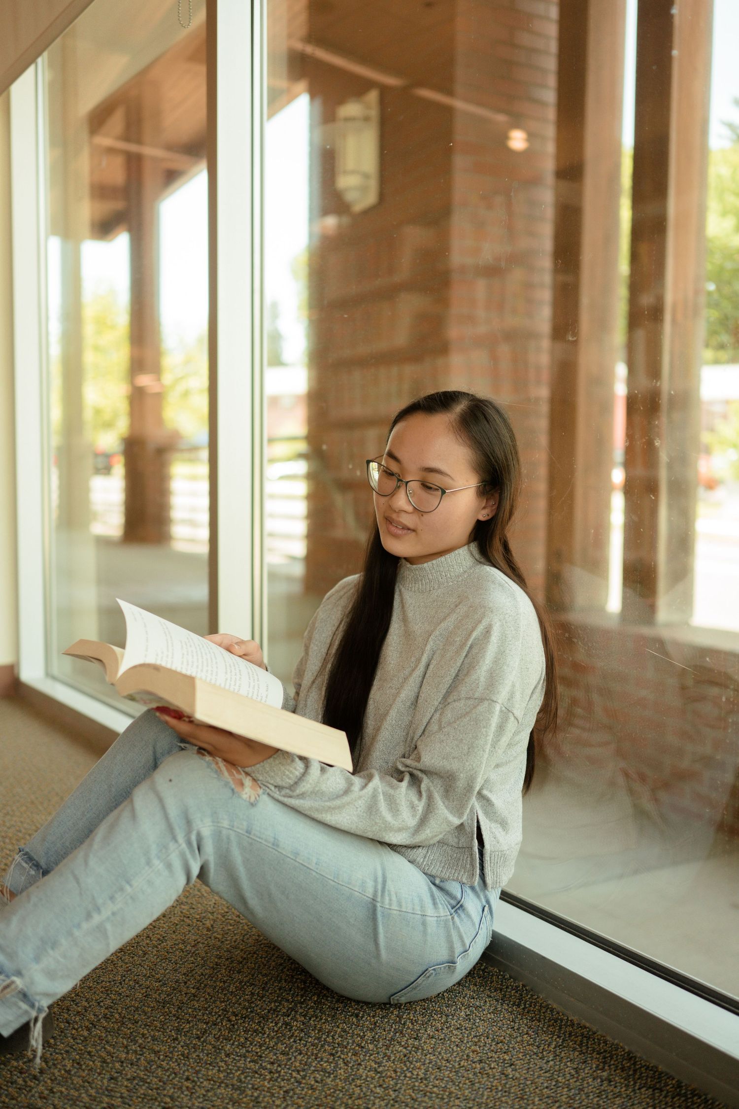 Student reading book while sitting by window in casual attire.