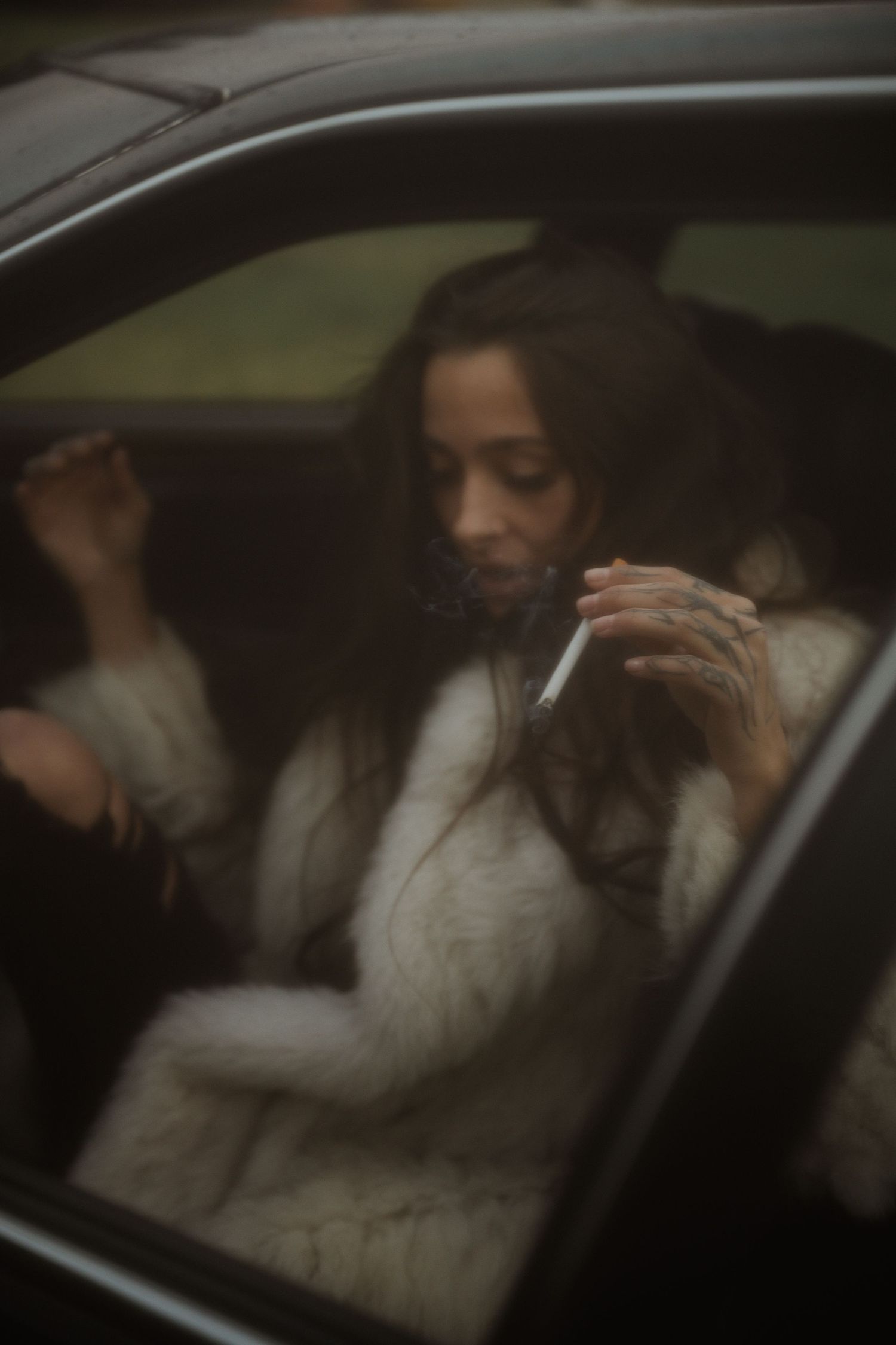 Through a car window, a figure in a white fuzzy sweater poses with long dark hair in a moody atmospheric setting.